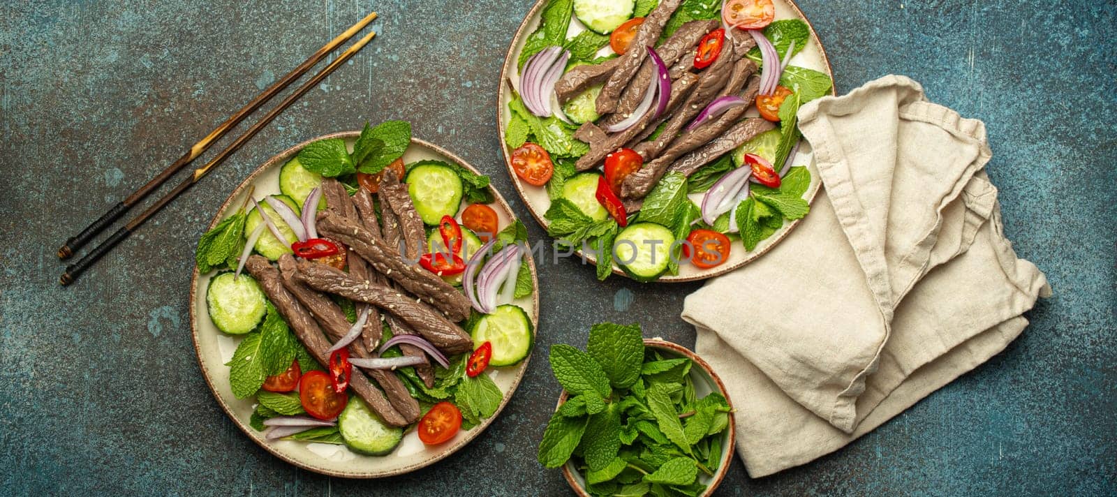 Two plates with traditional Thai beef salad with vegetables and mint top view served on rustic concrete background, healthy exotic asian meal.