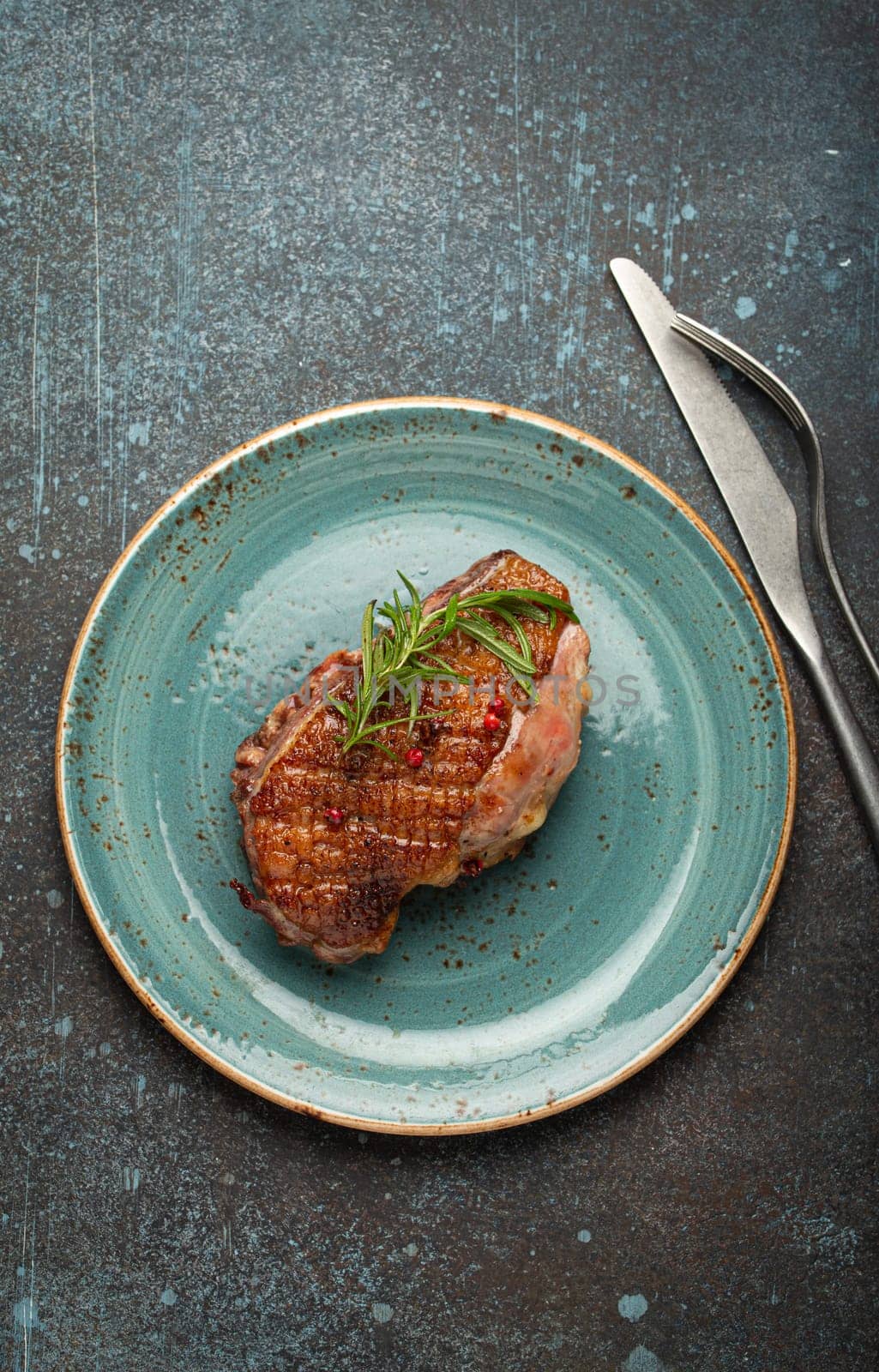 Delicious roasted duck breast fillet with golden crispy skin, with pepper and rosemary, top view on ceramic blue plate, rustic concrete rustic background.