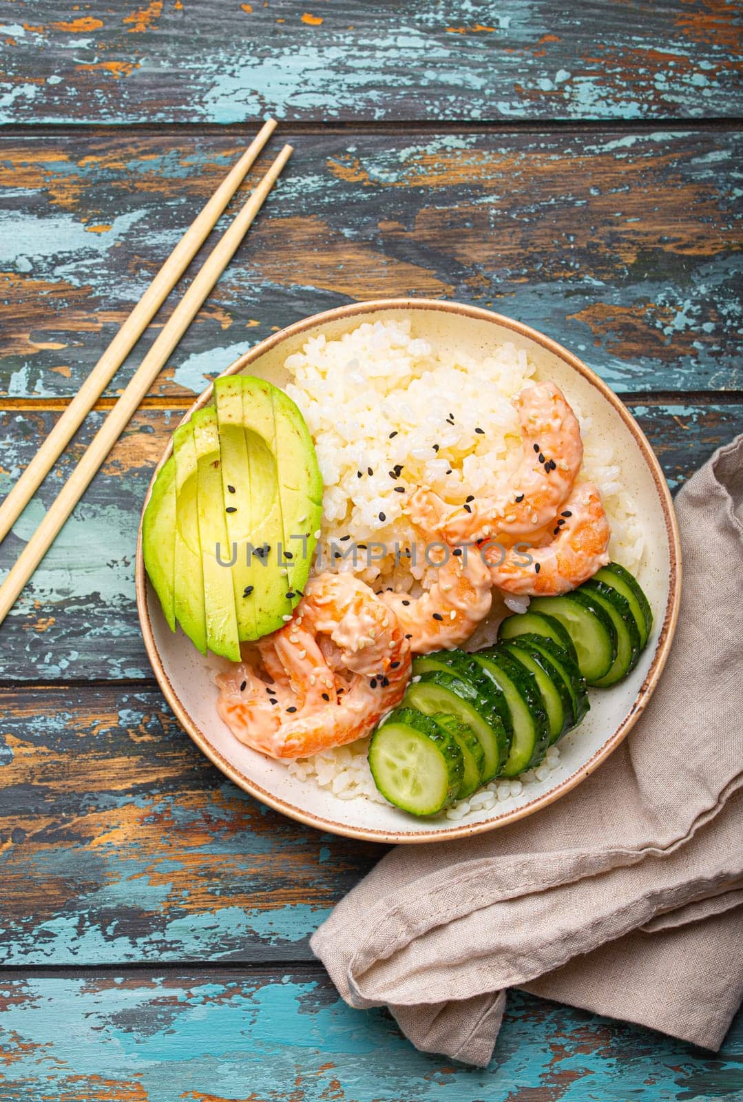 White ceramic bowl with rice, shrimps, avocado, vegetables and sesame seeds and chopsticks on colourful rustic wooden background top view. Healthy asian style poke bowl.