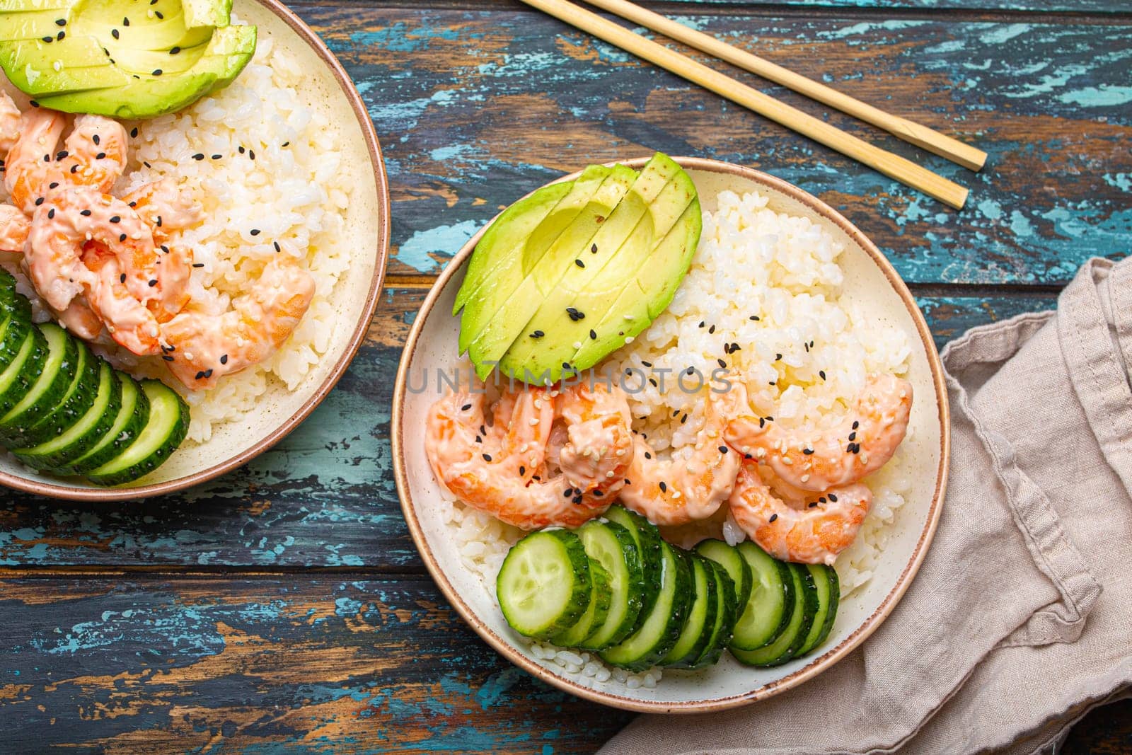 Two white ceramic bowls with rice, shrimps, avocado, vegetables and sesame seeds and chopsticks on colourful rustic wooden background top view. Healthy asian style poke bowl by its_al_dente