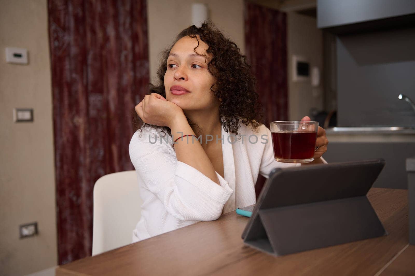 Relaxed young pretty woman in white bathrobe, holding cup of tea, dreamily looking aside, sitting t kitchen table in front of digital table in modern home interior. People. Communication. Internet. by artgf