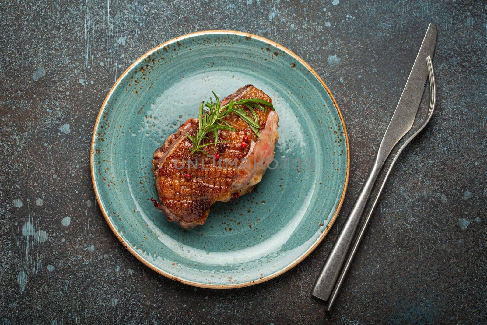 Delicious roasted duck breast fillet with golden crispy skin, with pepper and rosemary, top view on ceramic blue plate, rustic concrete rustic background.