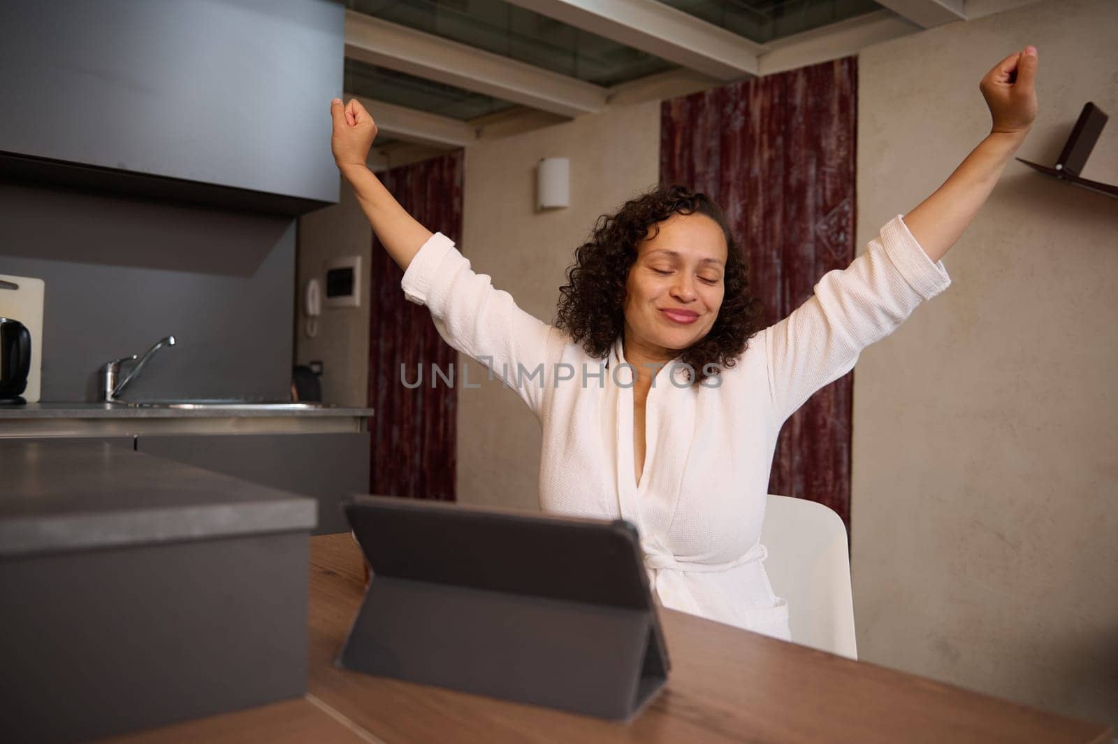 Charming young woman smiling stretching up after waking up in the morning, sitting at table with a digital tablet. People. Morning routine. Lifestyle. Online communication and business. Remote job