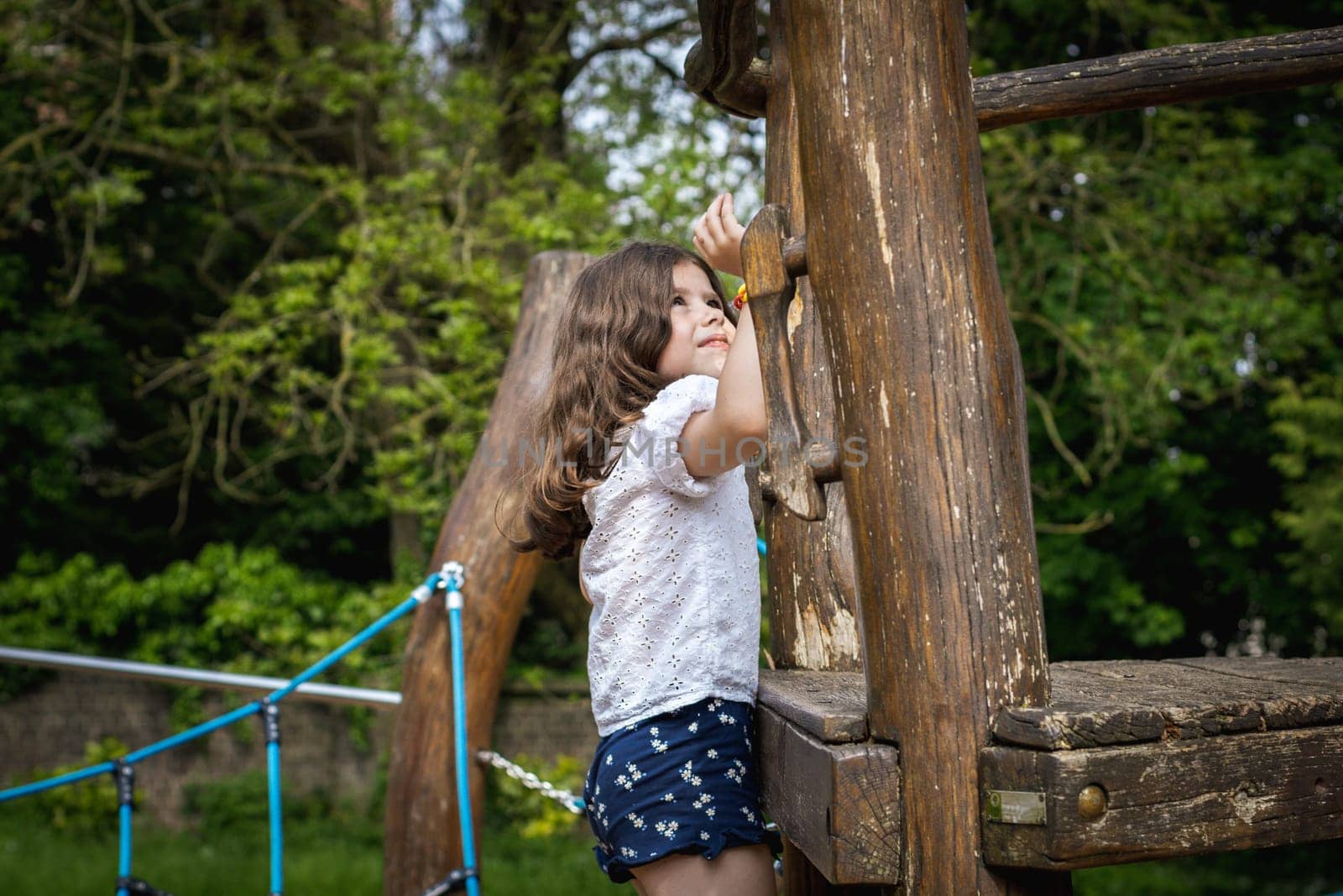 Portrait of one beautiful Caucasian brunette girl climbing on a wooden walkway to a rope swing on a playground on a summer day in a park on a playground, close-up side view.