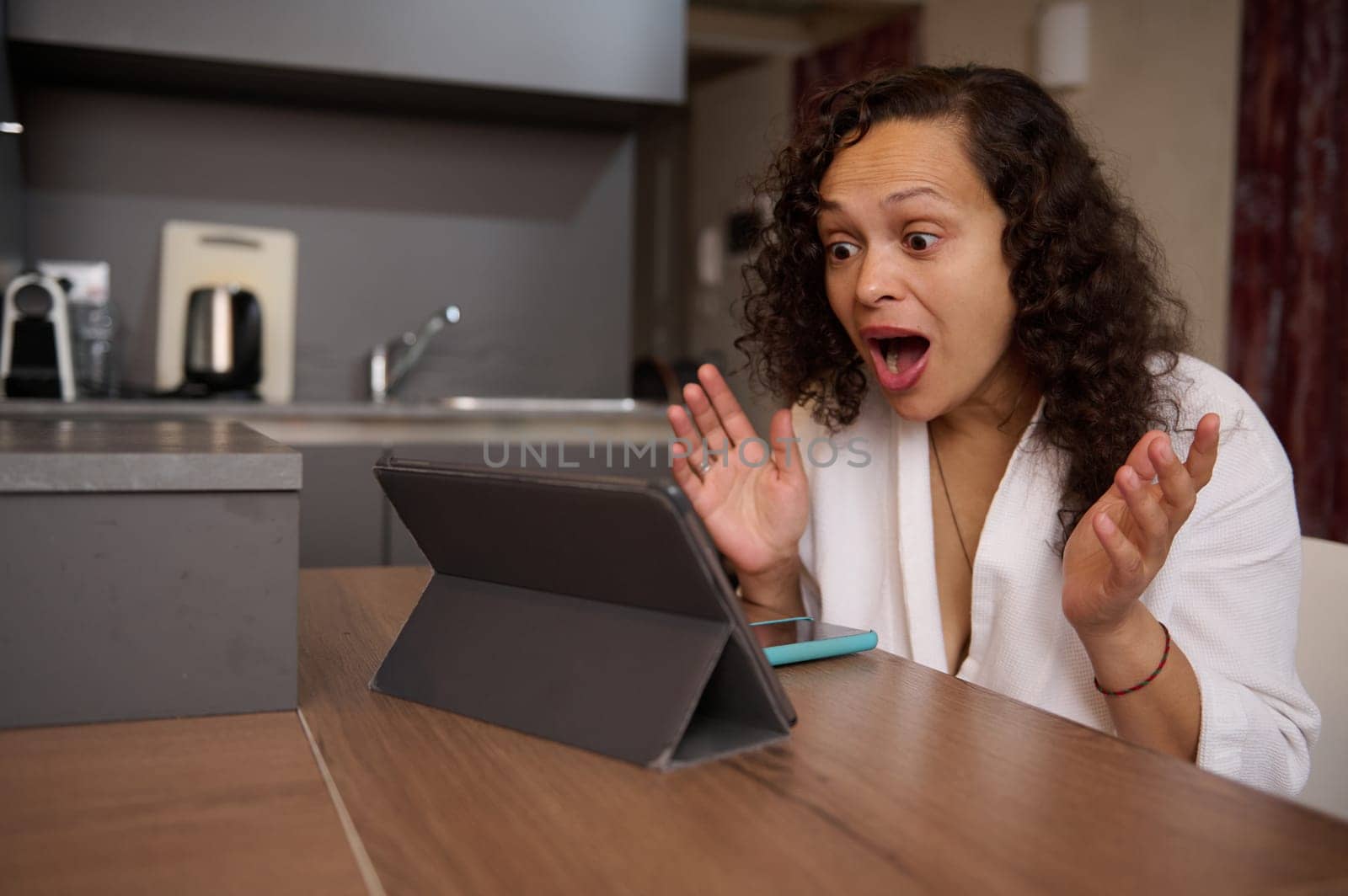 Emotional portrait of a young woman in white bathrobe, expressing amazement astonishment looking at the digital tablet, sitting at kitchen table at home. by artgf