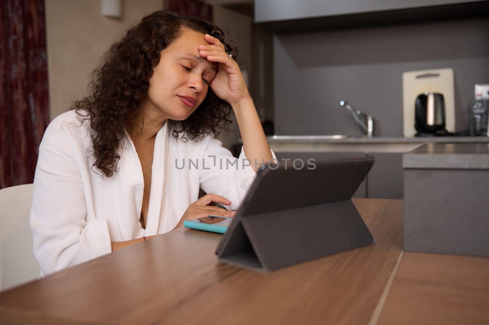 Tired business woman in white bathrobe at home, working online on tablet, reading email with bad news, suffering from burnout, sitting at kitchen table. Young female boss dealing with work problem by artgf