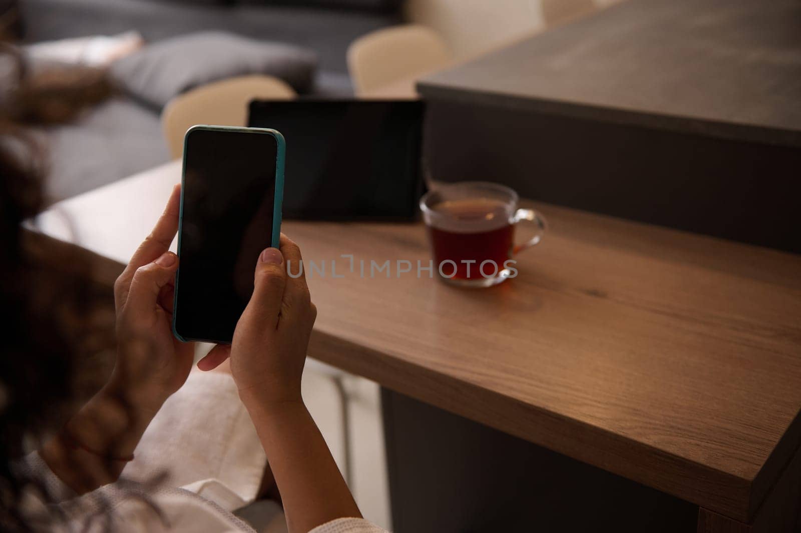 Close-up rear view of a woman sitting at table at home, texting on mobile phone with black mockup digital screen. Copy space for advertising text or mobile application by artgf