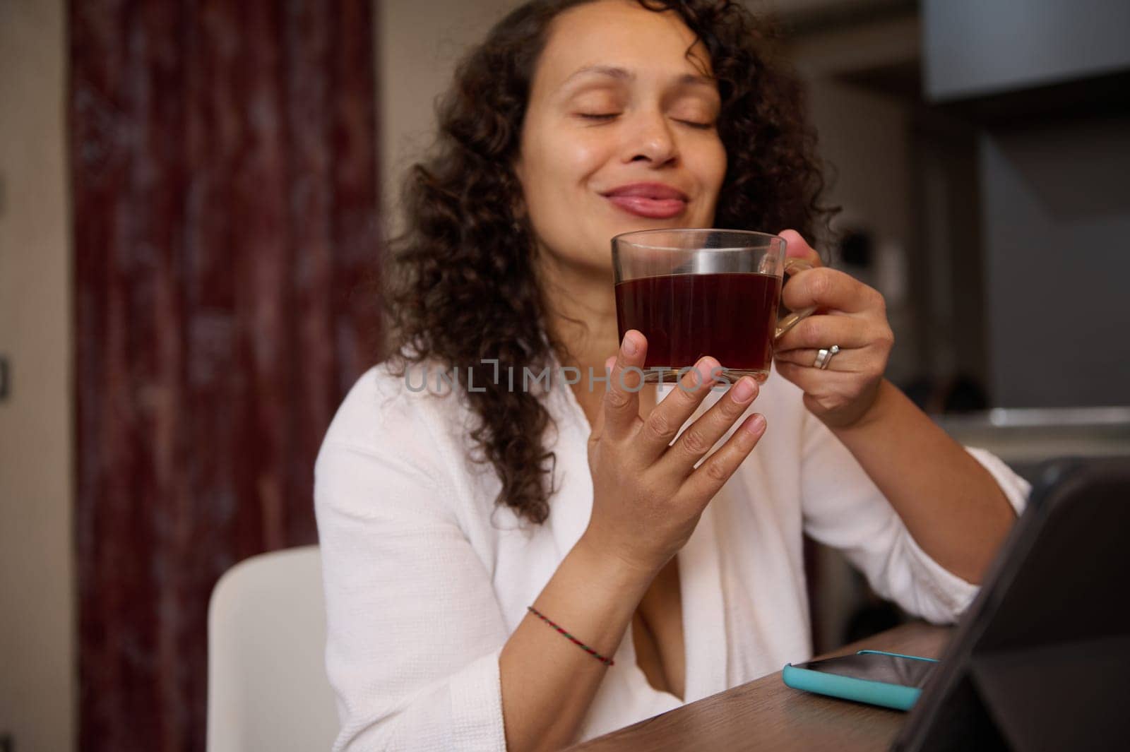 Happy curly haired multi ethnic woman in white waffle bathrobe, smiles relaxing over up of hot tea in home interior. Focus on a glass cup of hot healing herbal drink in women's hands by artgf
