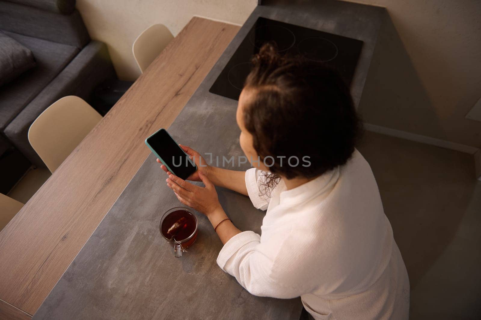 Overhead view of young adult woman holding a smartphone with blank black mockup touch screen, making online payment, shopping, paying bills, sitting at kitchen table in the home office. Copy ad space