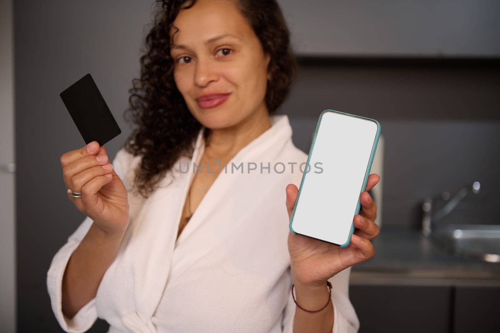 Young woman holds and shows at camera blank black credit card and smartphone with white empty mockup digital screen, makes online payments, transfers money via internet mobile banking or application by artgf