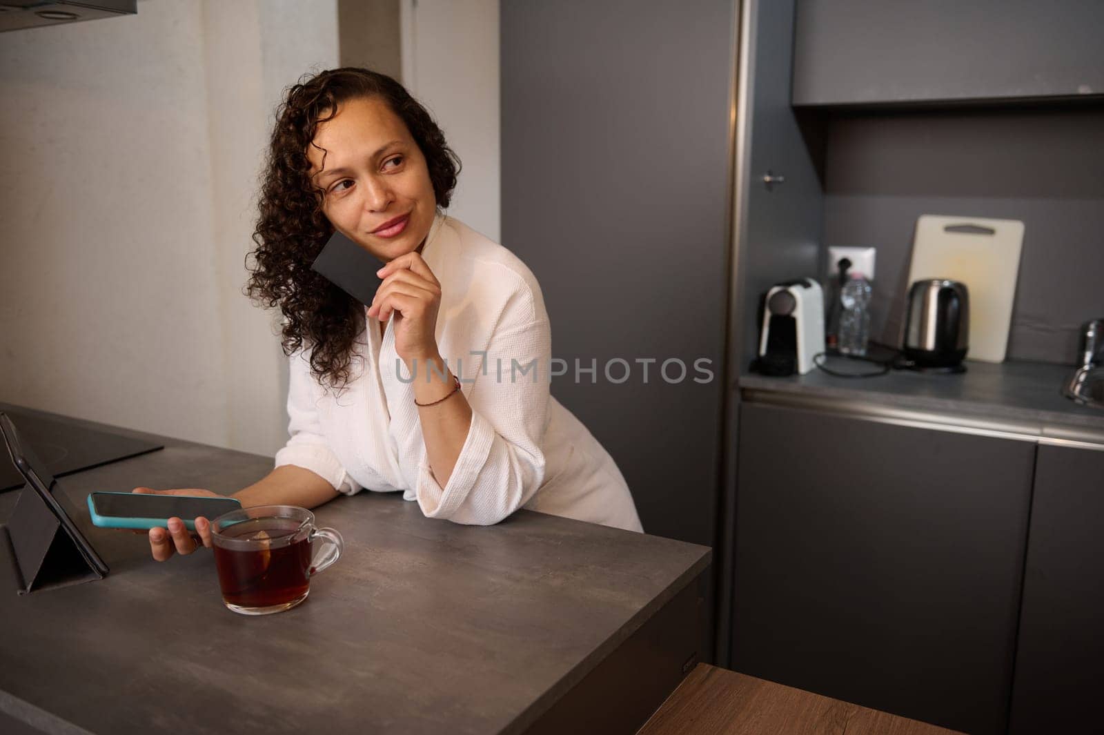 Middle aged beautiful confident woman holding bank credit card, using smartphone, online shopping, paying bills, receiving or making payment via mobile internet banking, standing at table at home