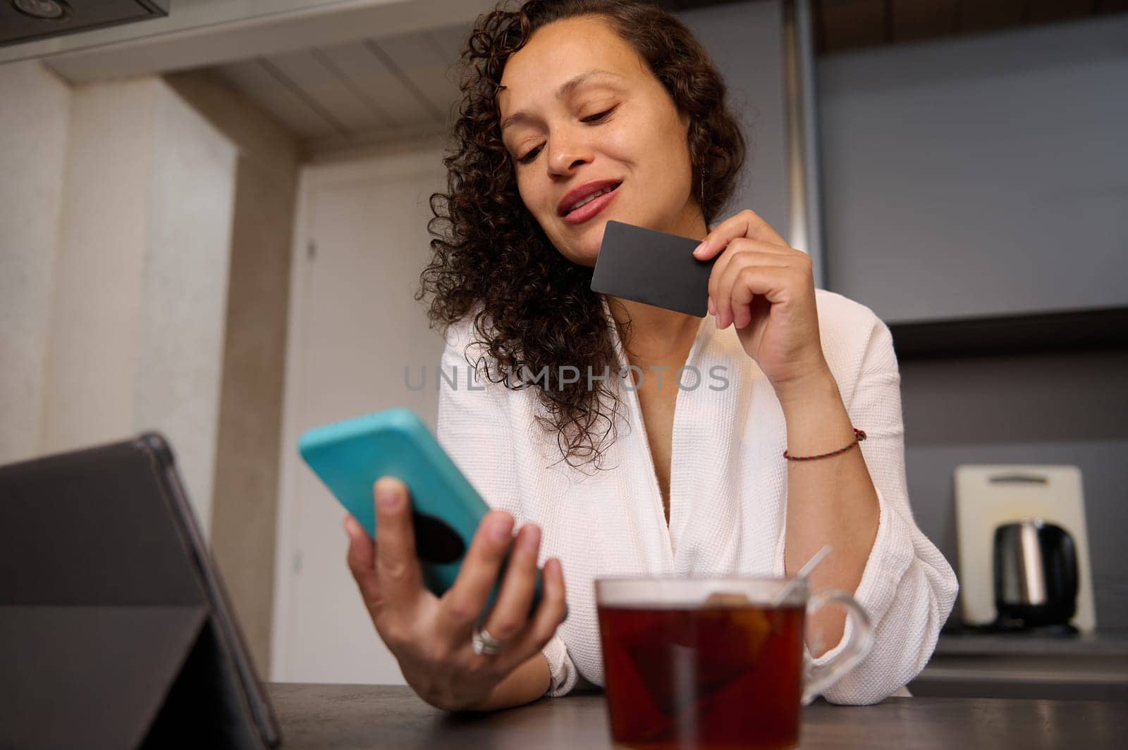 Smiling woman at home kitchen, using mobile phone and online shopping, paying bills online via internet mobile banking application by artgf