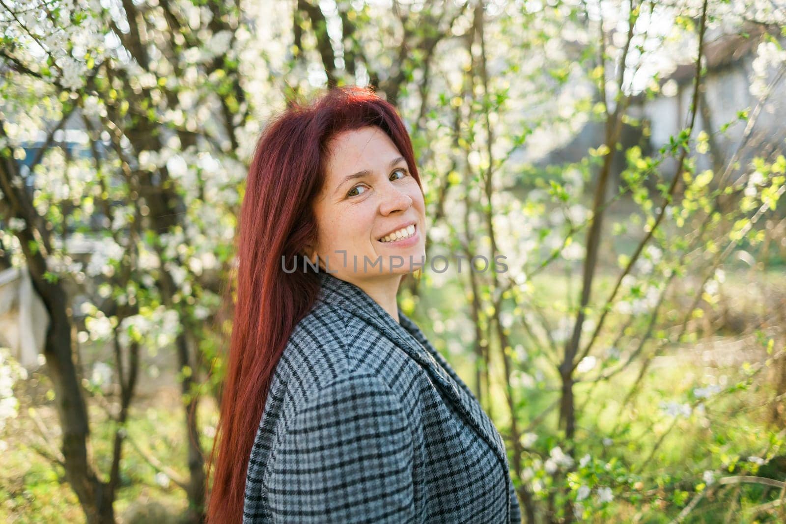 Beautiful red-haired woman enjoying smell in a flowering blooming spring garden. Spring blossom. Copy space and empty place for advertising text by Satura86