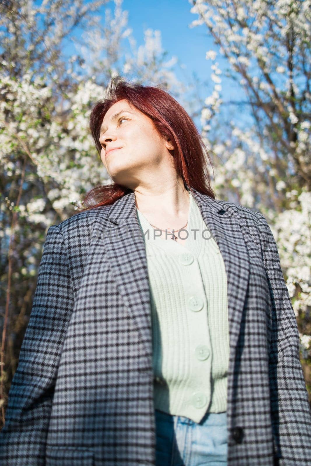Beautiful red-haired woman enjoying smell in a flowering blooming spring garden. Spring blossom. Copy space and empty place for advertising text by Satura86