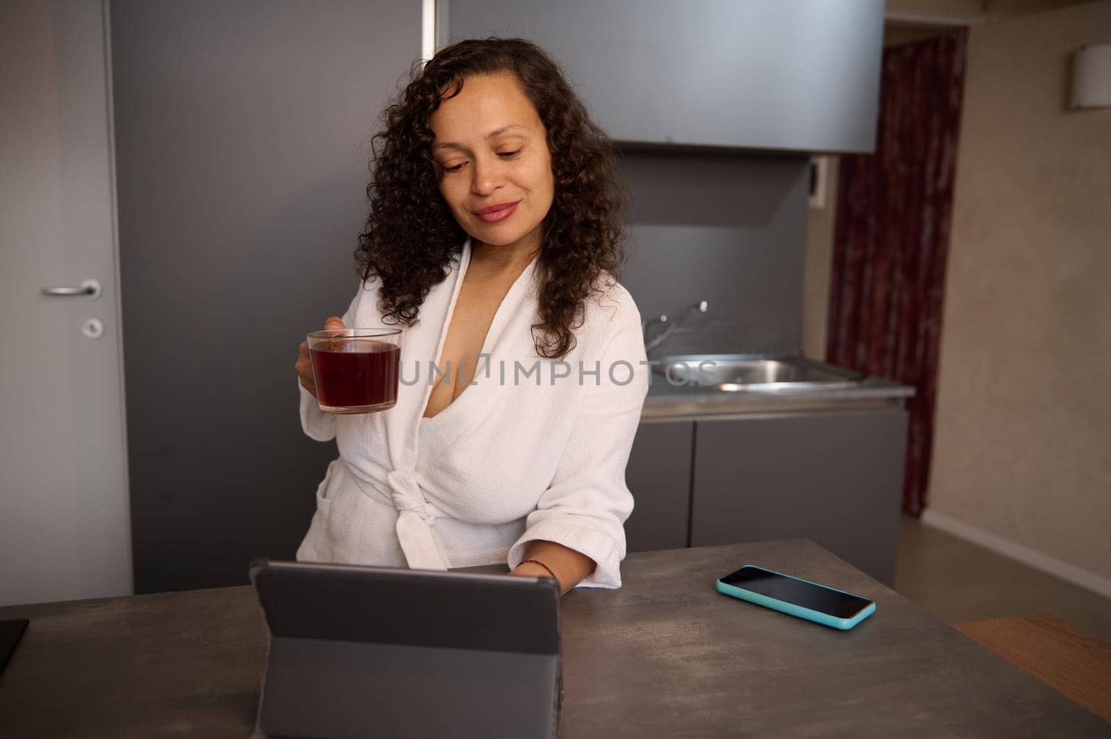 Portrait of relaxed young multi ethnic woman freelancer holding a cup of hot tea, reading news on her digital tablet, checking social media content, standing at kitchen counter in modern home interior