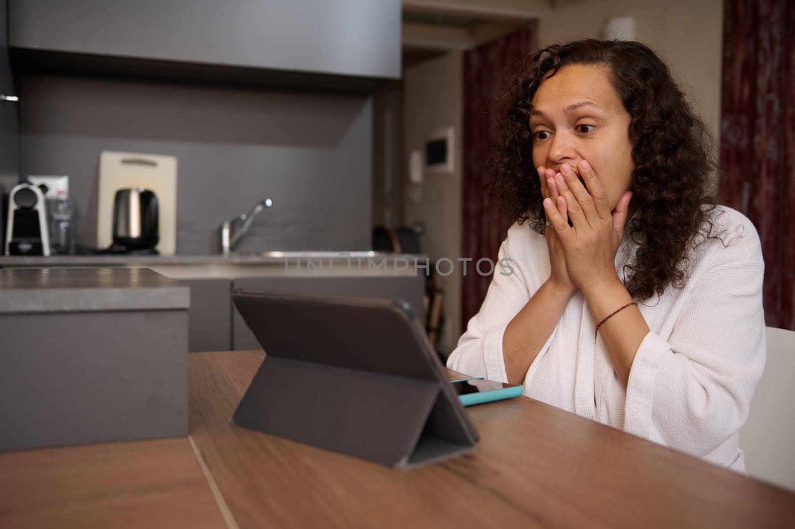 Emotional young woman in white bathrobe, using digital tablet at home, expressing worry and stupefaction reading news, sitting at kitchen table at home. Brunette looks afraid while watches scary movie