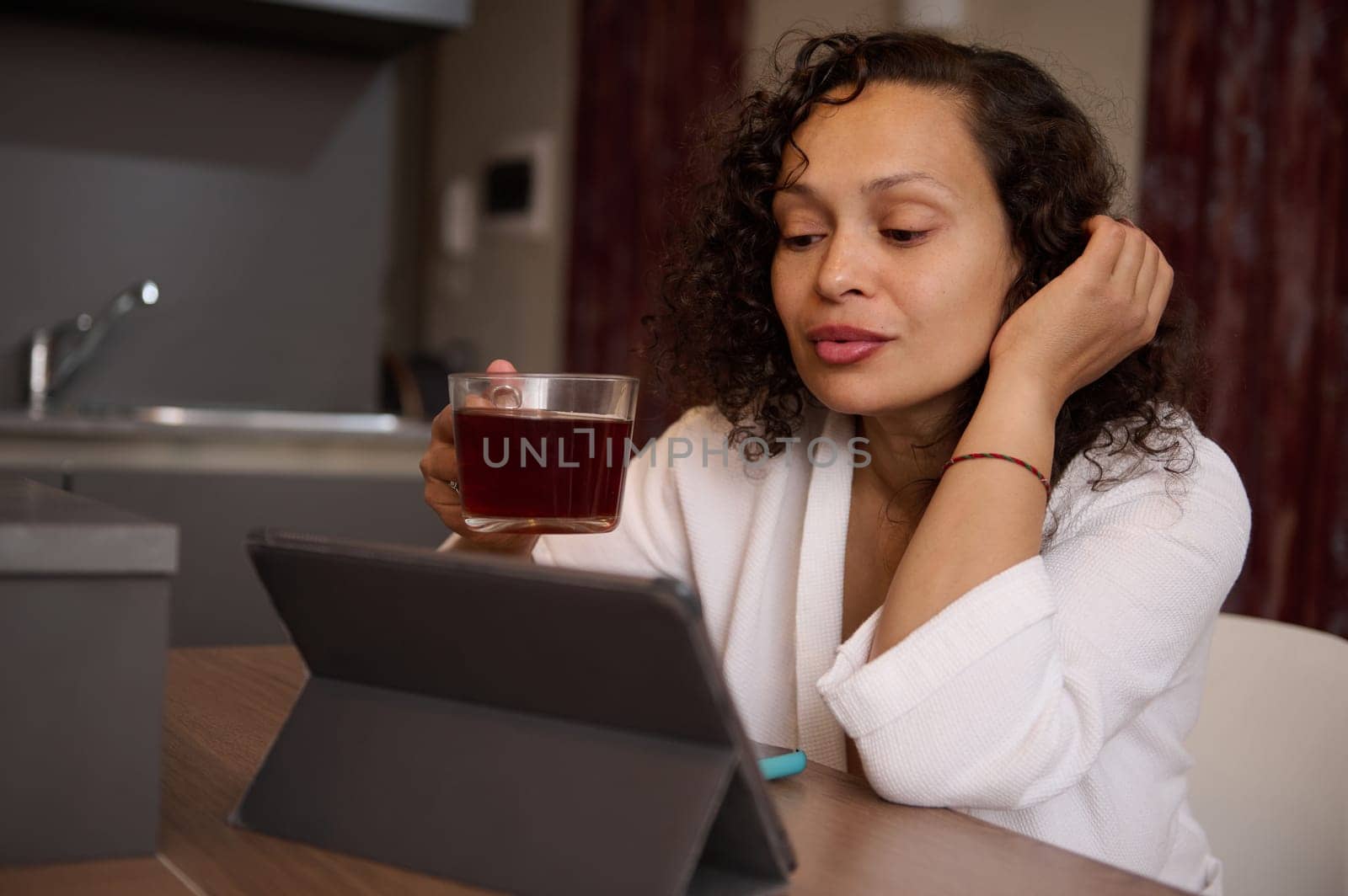 Attractive delightful woman reading news on digital tablet, browsing web sites, sitting at kitchen table over cup of tea by artgf