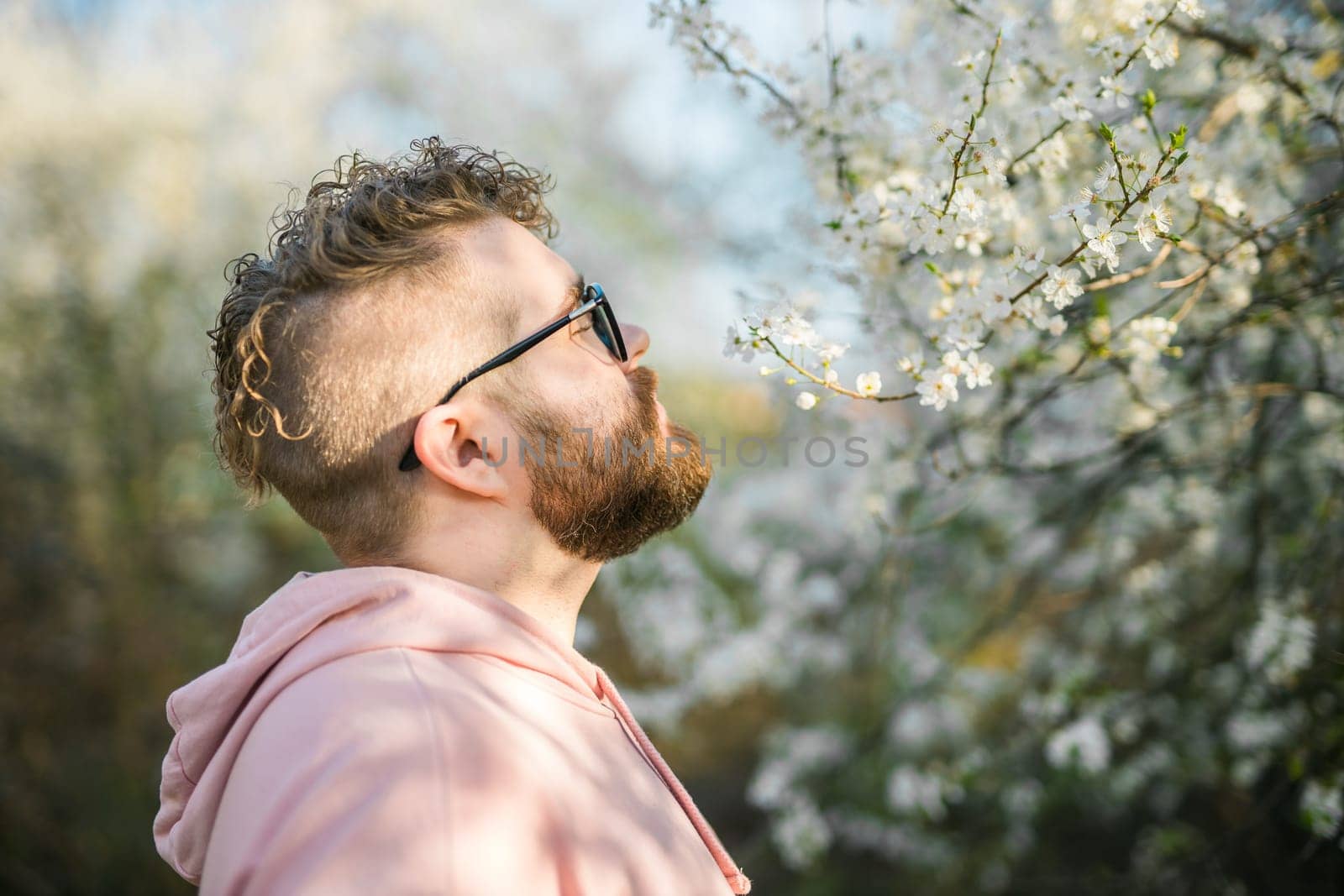 Portrait of curly millennial man inhales the fragrance of spring flowers of blooming jasmine or cherry tree. Spring time