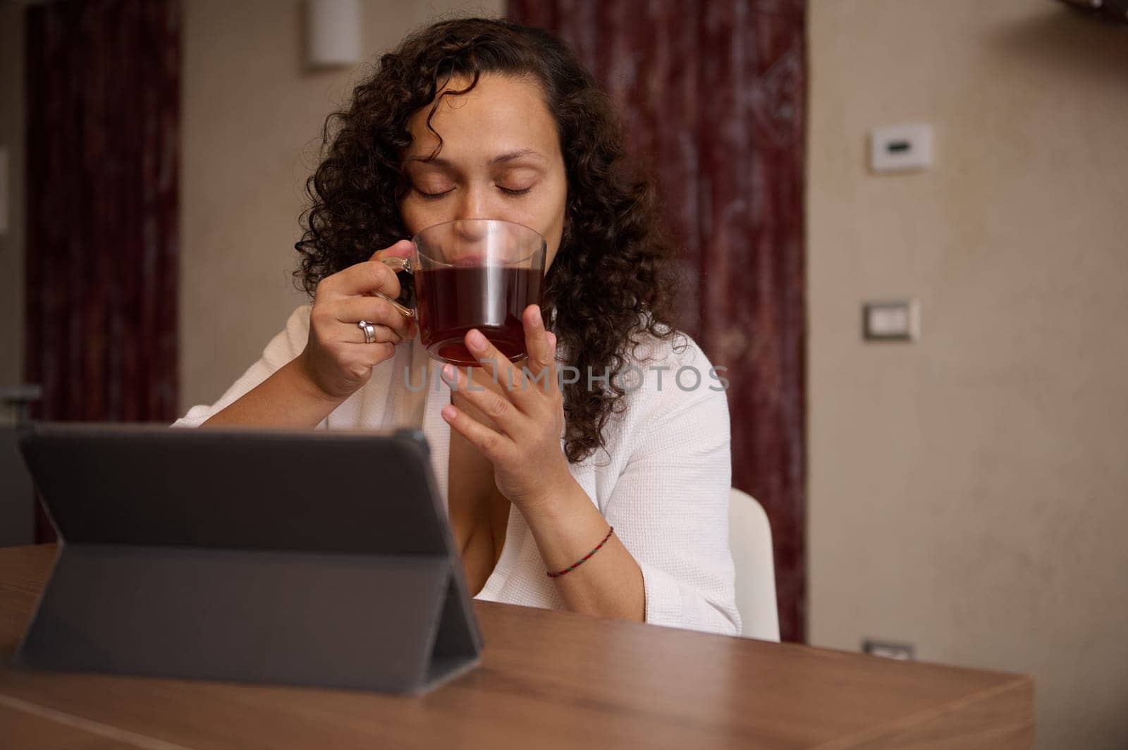 Beautiful multi ethnic middle aged woman in white bathrobe, drinking hot tea, sitting at digital tablet at home interior by artgf