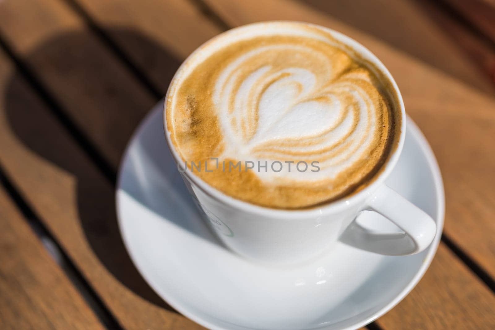 Coffee on wooden background. Cup of cappuccino with latte art on brown table with sunbeam. Fresh morning coffee with delicious milk foam. by Satura86