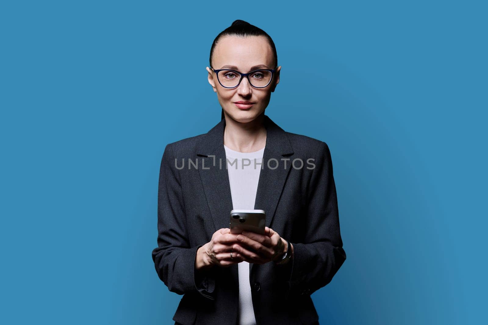 Adult smiling woman holding smartphone on blue background. 30s female looking at camera with phone in hands. Technologies mobile applications internet work business leisure communication