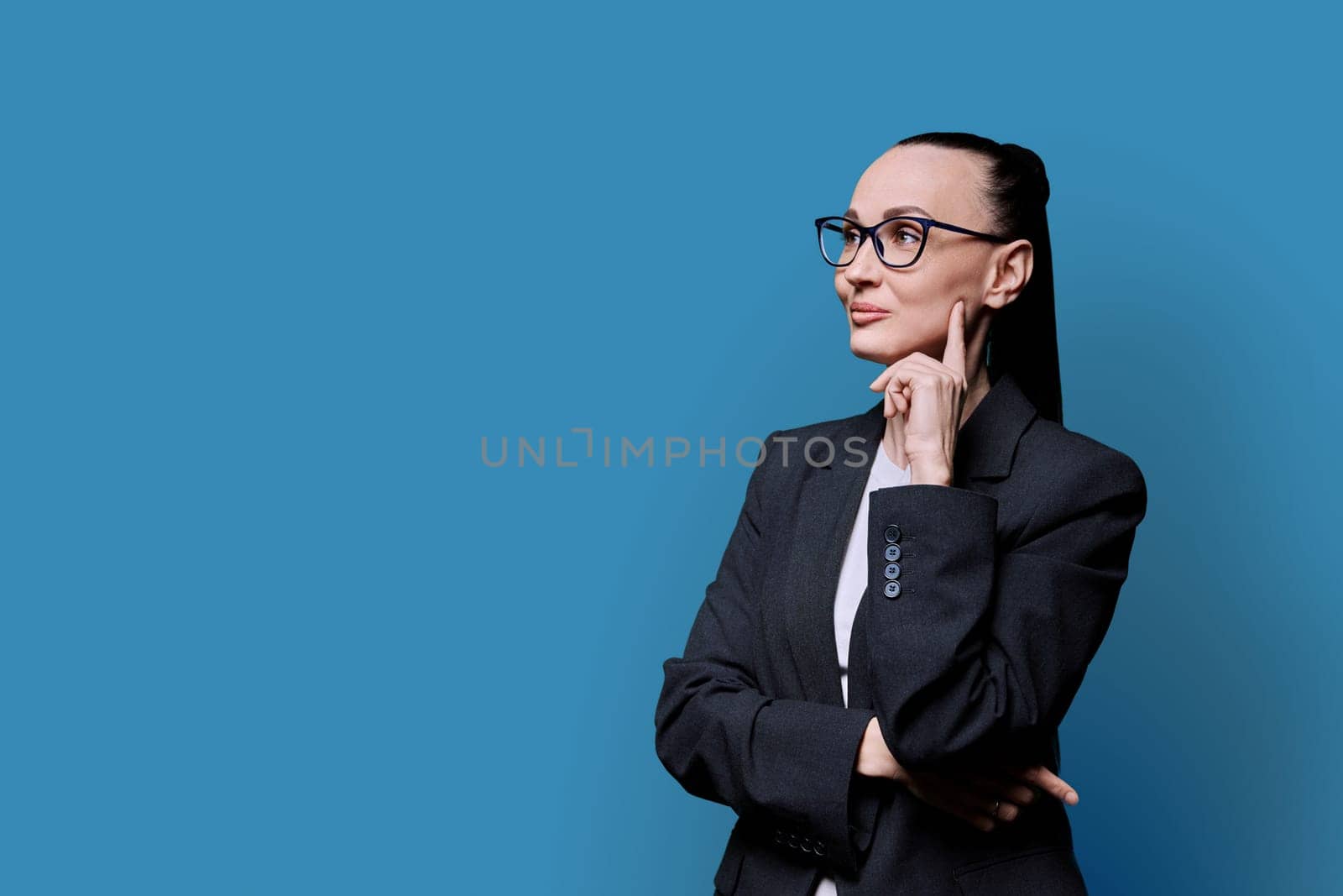 Portrait of thinking serious 30s business woman on blue studio background. Confident female in glasses suit looking to side at space for image text copy space. Business work teaching job career people