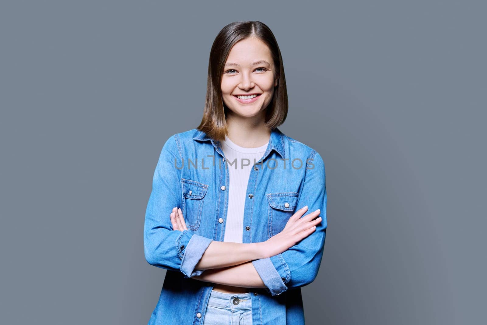 Young beautiful confident woman with crossed arms on gray studio background. Smiling attractive 20s female in denim looking at camera. Lifestyle, fashion beauty style, youth business work education