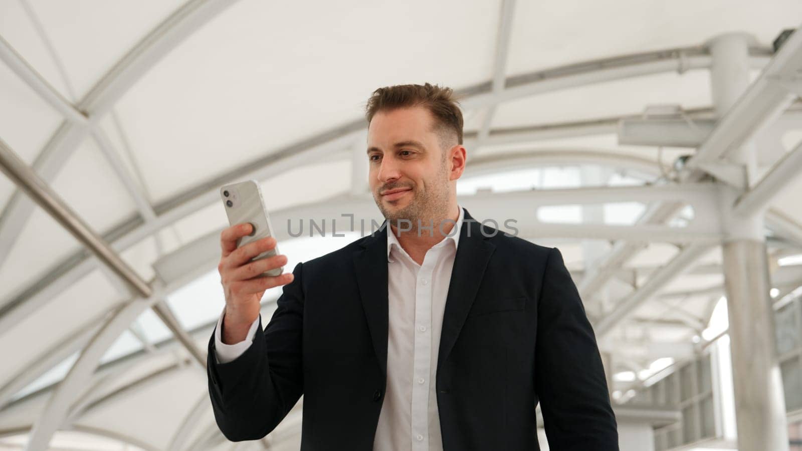 Skilled businessman looking or checking his mobile phone while walking at bridge for going to workplace. Happy manager using telephone to send massage to marketing team and receive good news. Urbane.