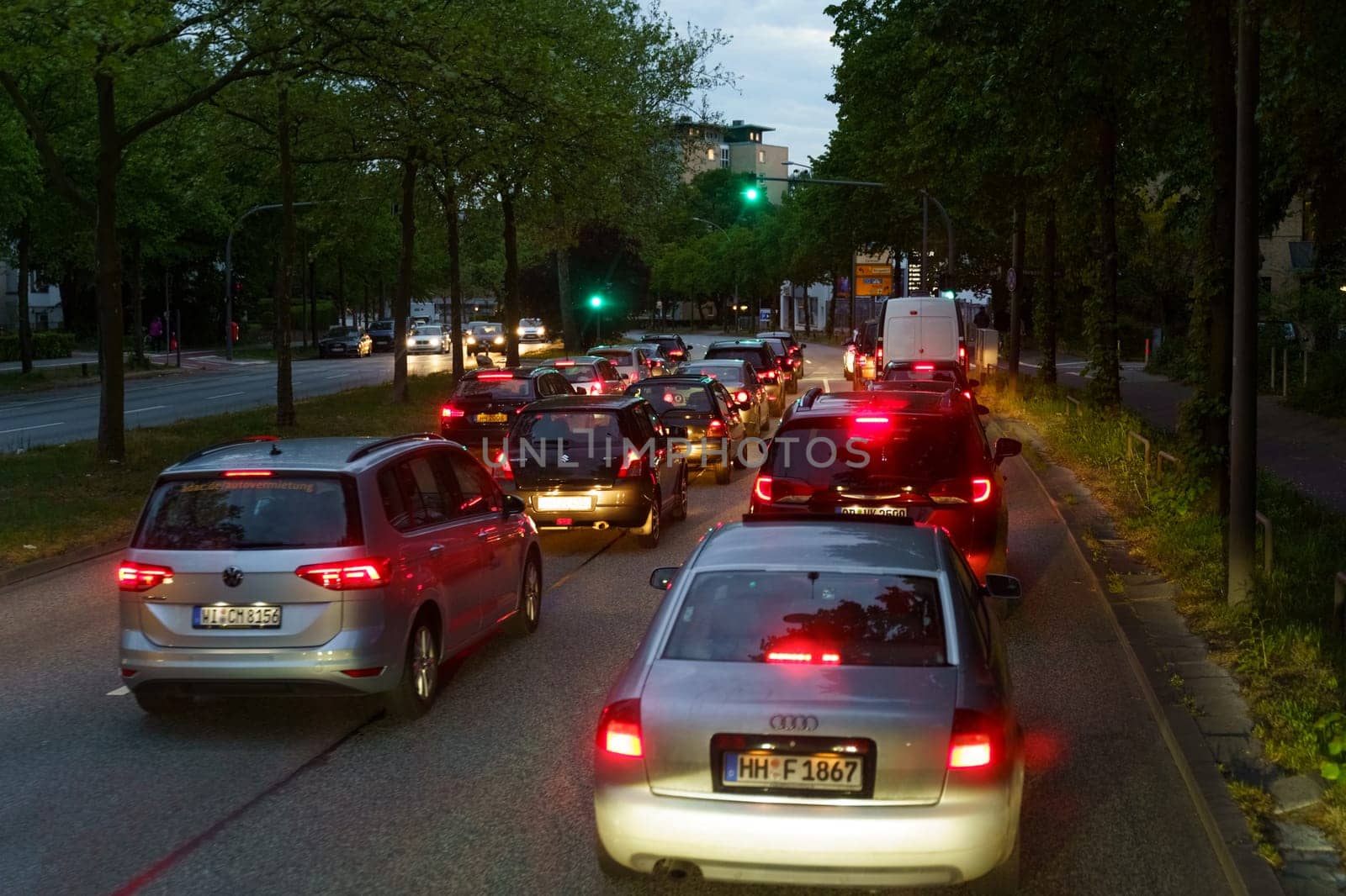 Hamburg, Germany - May 19, 2023: Vehicles line up at a traffic light on a city street during twilight, with the glow of red tail lights and street lamps illuminating the scene.