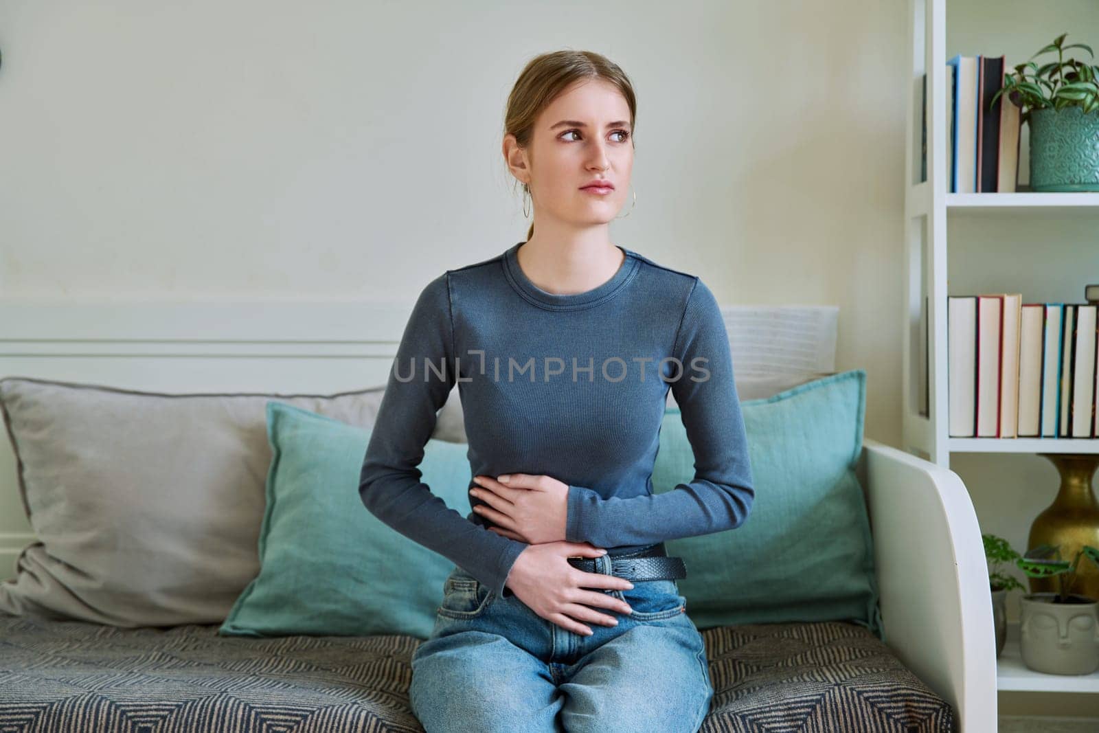 Young teenage female experiencing abdominal pain sitting at home. Stomach, menstrual cramps, gastrointestinal tract diseases, inflammatory processes in abdominal organs, kidneys, pelvis, youth, health