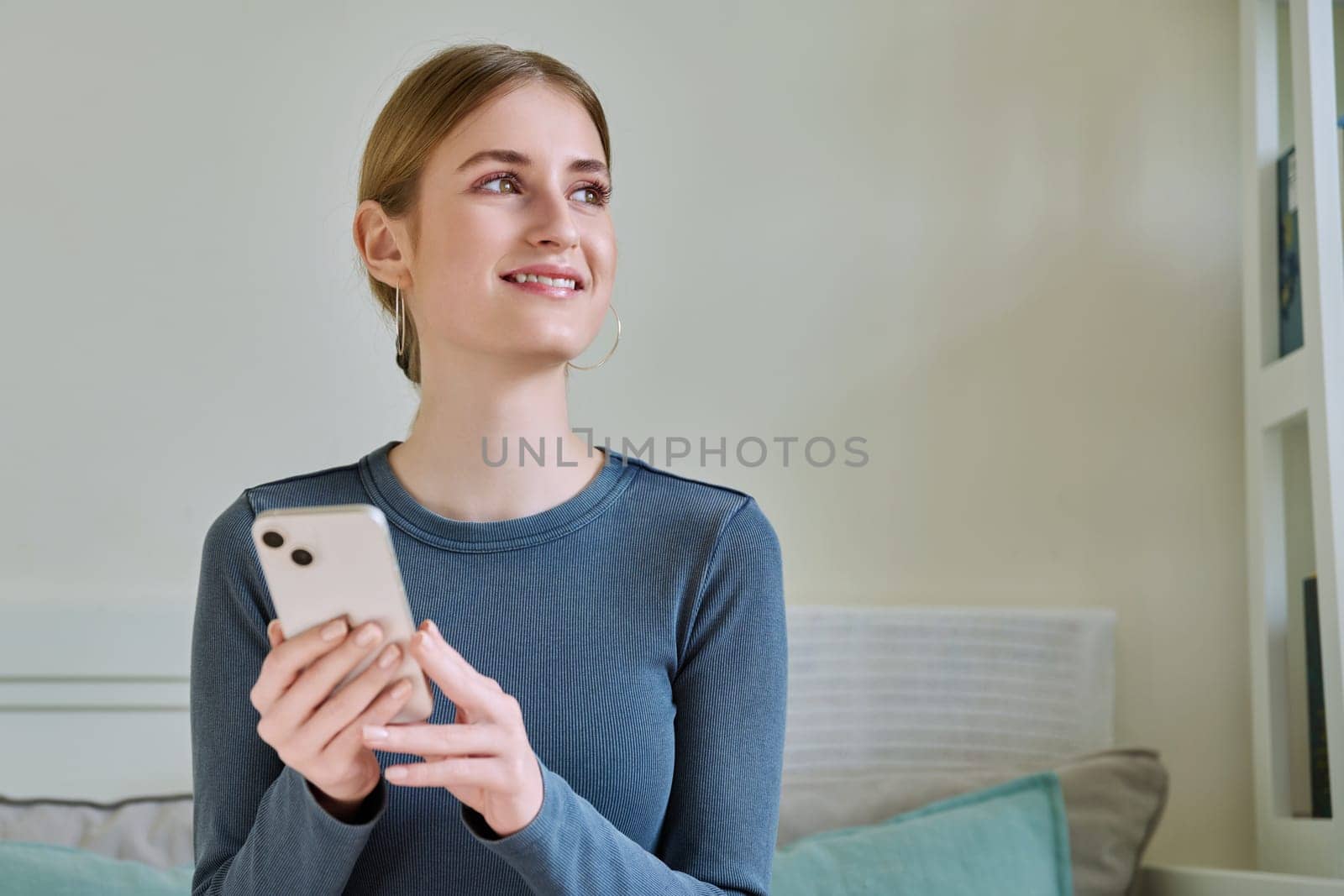 Happy smiling female teenager using smartphone, sitting on couch at home, girl 16,17, 18 years old texting reading messages. Modern digital technologies for communication, leisure, learning, shopping