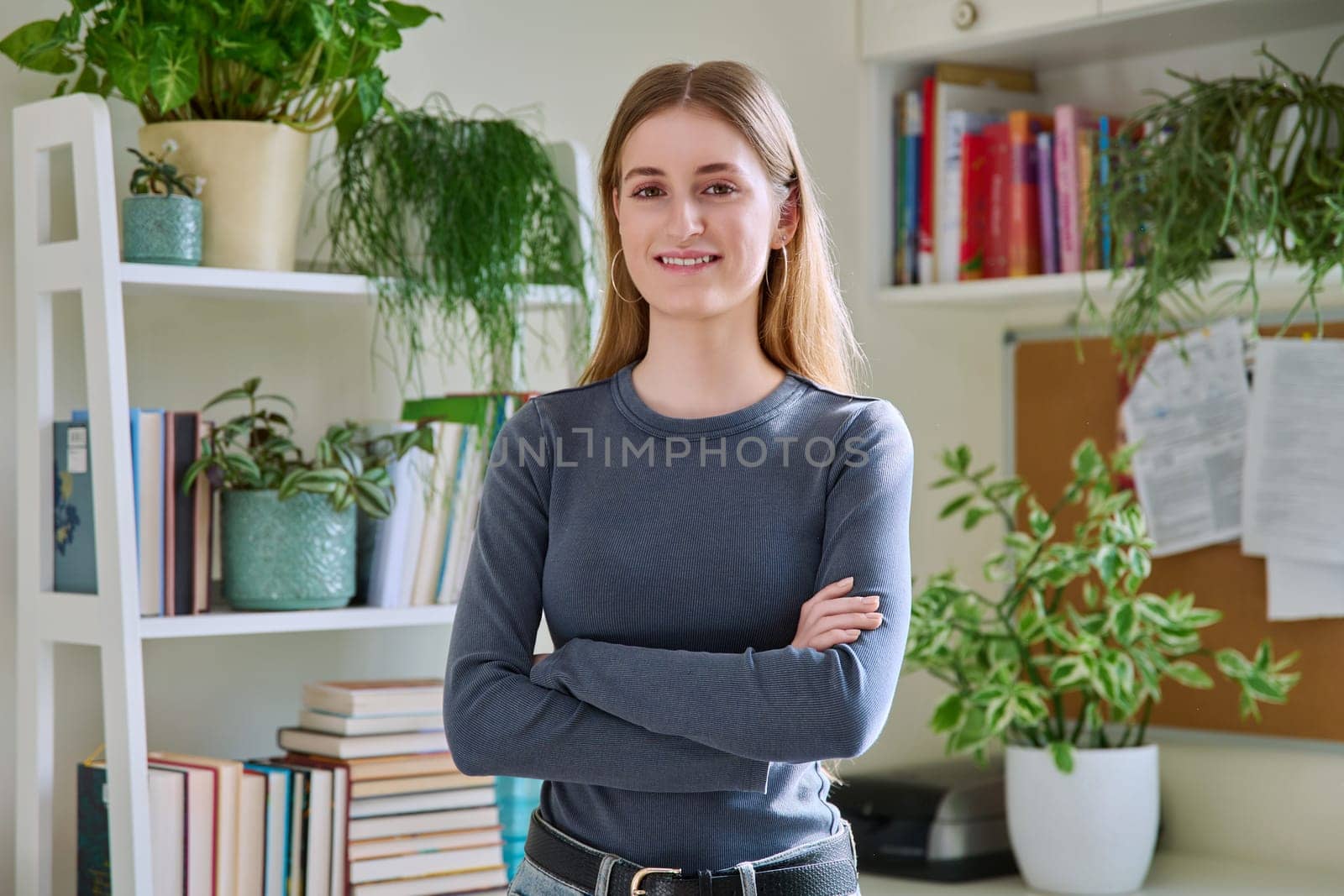 Portrait of female teenager 16-18 years old, smiling confident looking at camera with crossed arms in home. Beautiful blonde girl with long straight hair. Youth lifestyle beauty fashion