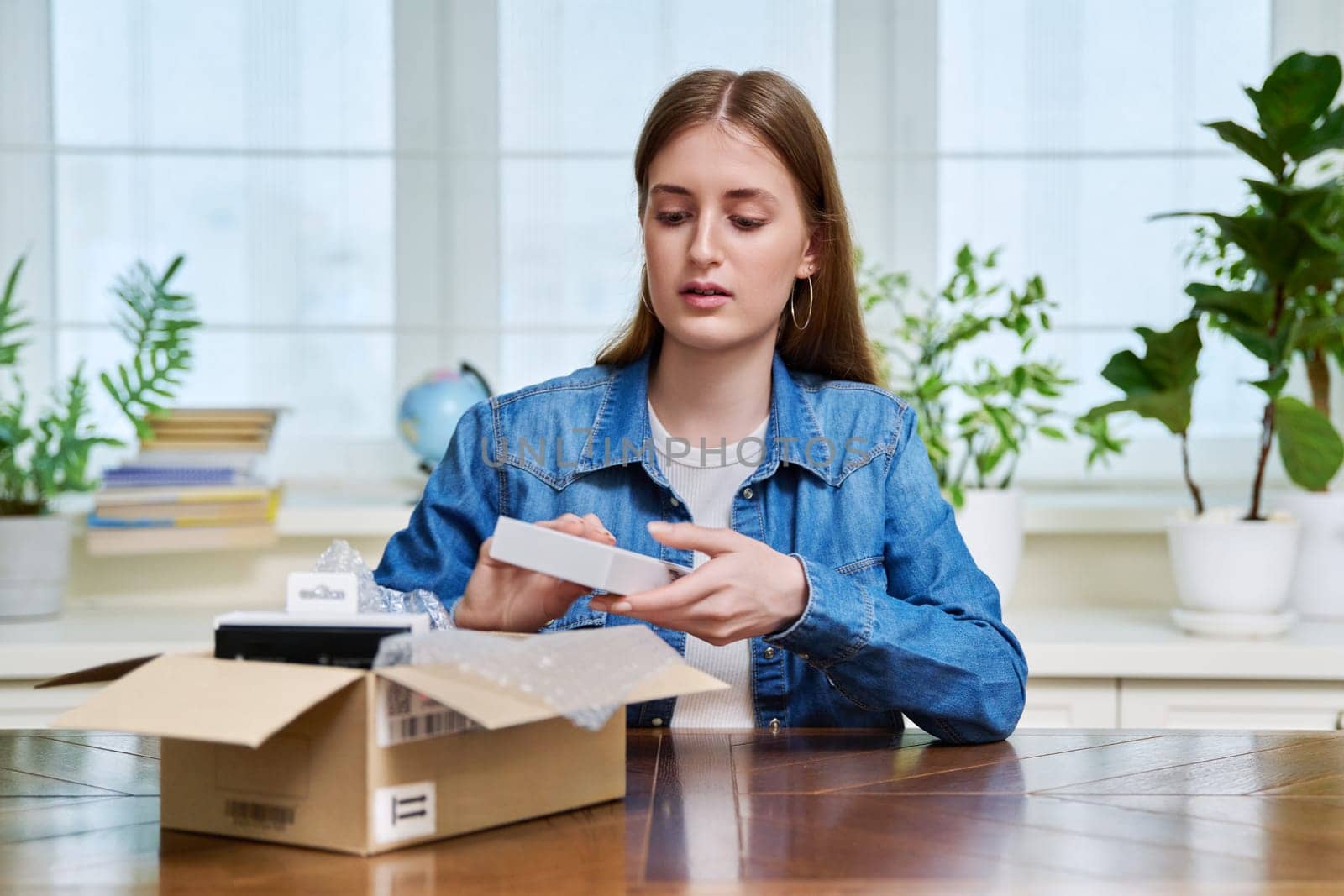 Satisfied young teen female shopper consumer sits at home and unpacks cardboard box with online purchases. Teenager girl unpacking box with new smartphone, headphones. Delivery by mail, online store