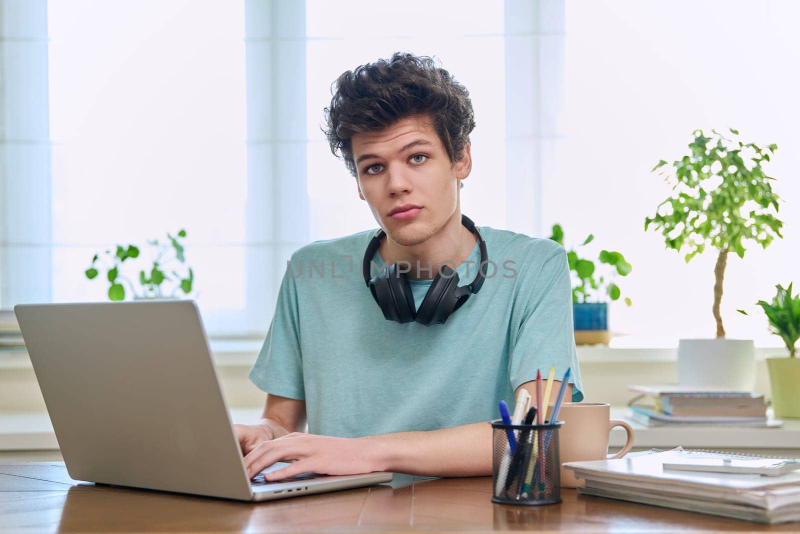 Young college student guy sitting at desk with laptop computer looking at camera. Handsome curly young male wearing headphones in home interior. Education, lifestyle, youth 19,20 years old