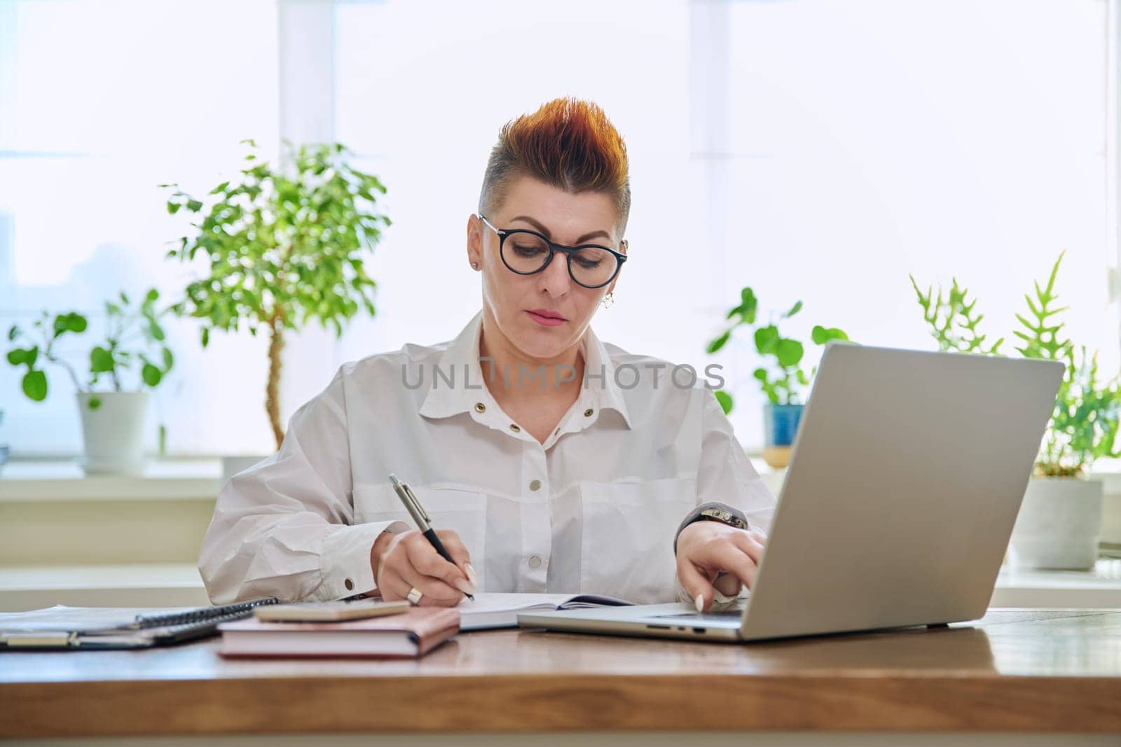 Middle-aged serious woman working at computer laptop in home office. Mature female sitting at desk typing and looking at monitor. Remote business, teaching, work, blogging, freelance