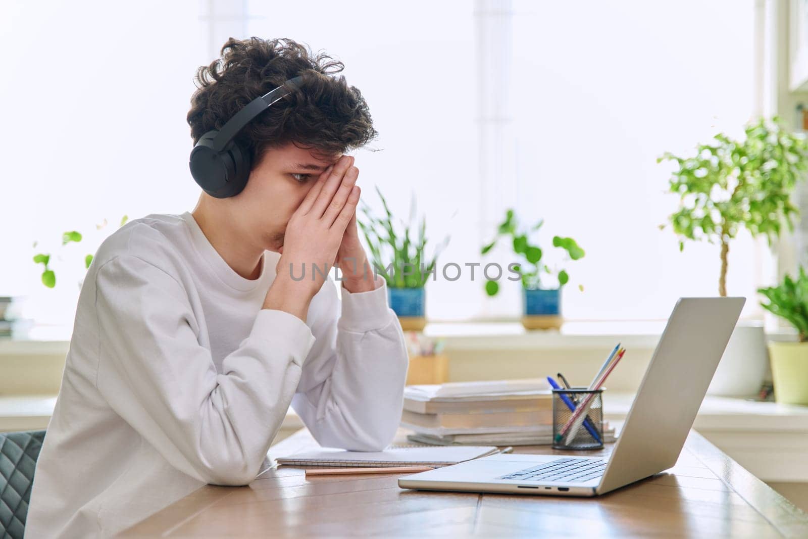 Sad upset young guy student in headphones sitting at home at desk with computer laptop, touching head with hands. Problems headaches troubles in college difficulties in study mental stress depression