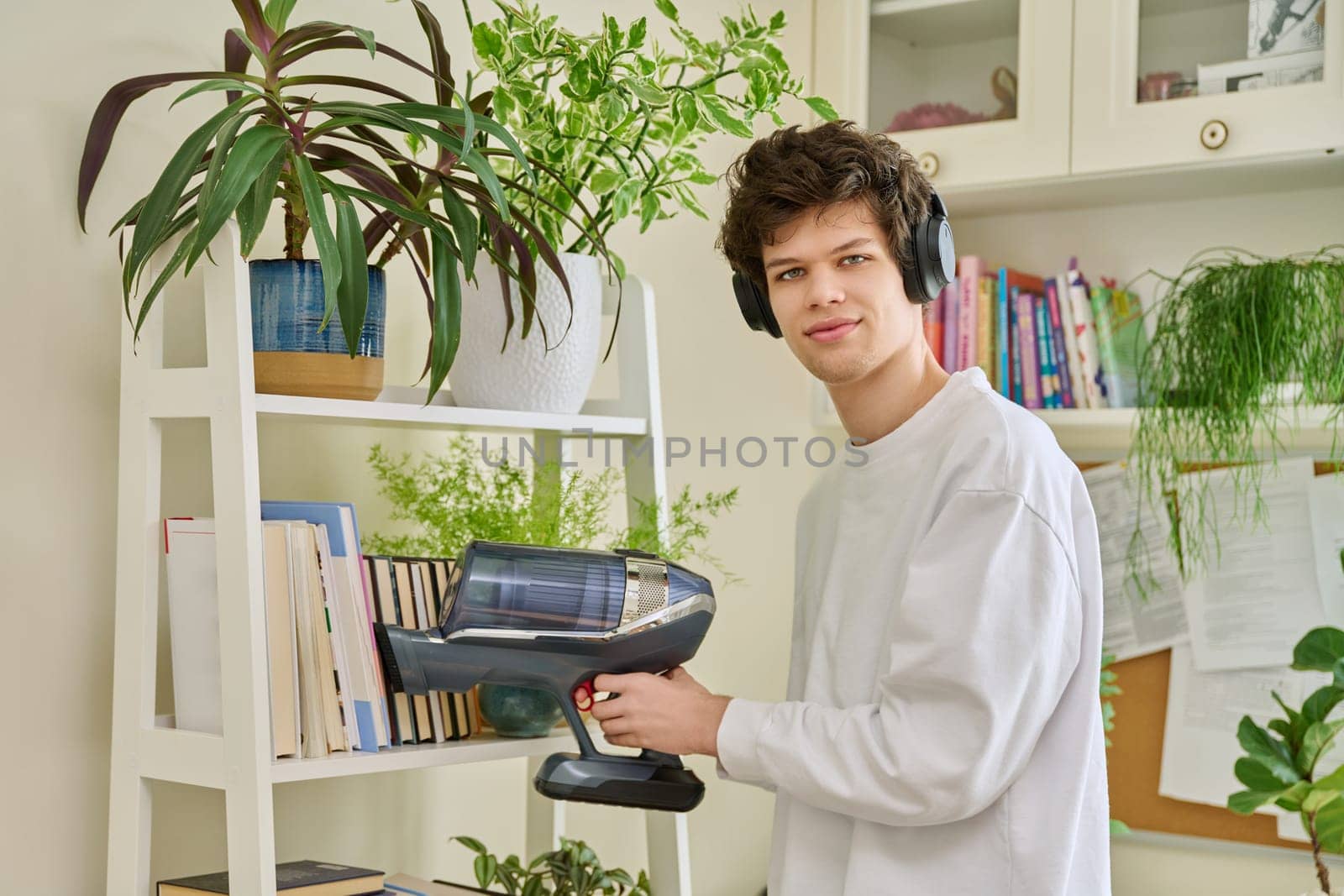 Handsome young man wearing headphones vacuums bookshelves at home. Cleaning, hygiene, housekeeping, housework, housecleaning, youth concept