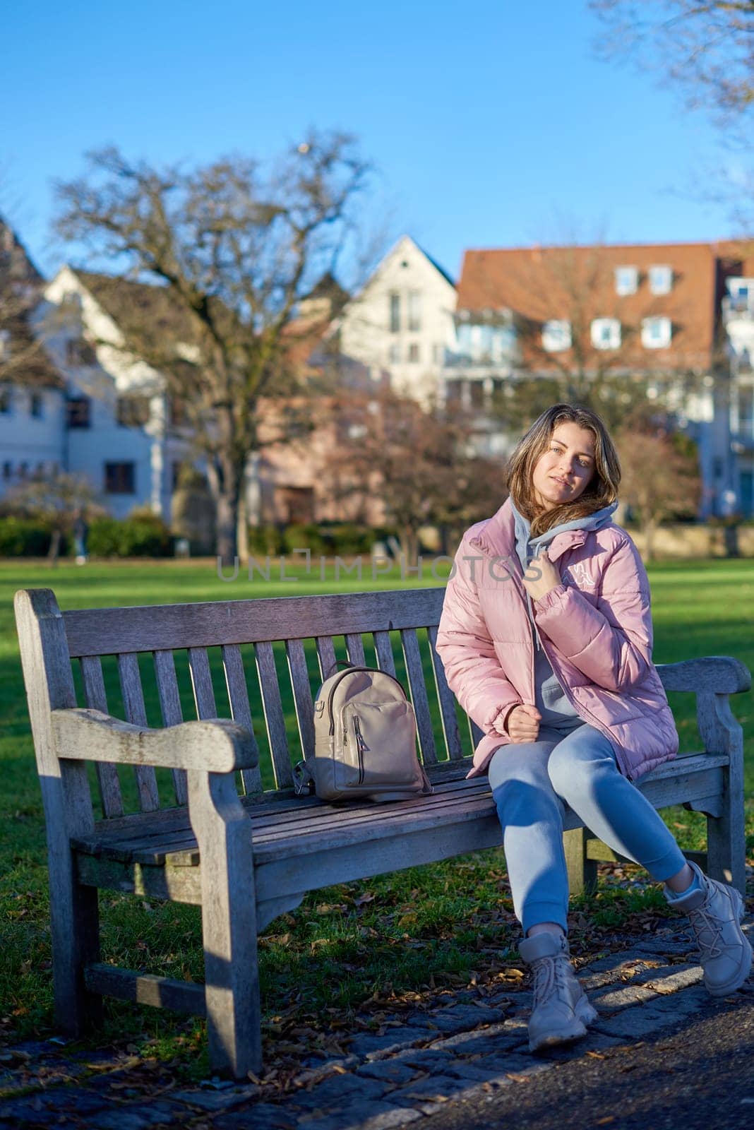 Beautiful Girl in Pink Jacket Relaxing on a Park Bench in the Background of an Old European Town. by Andrii_Ko