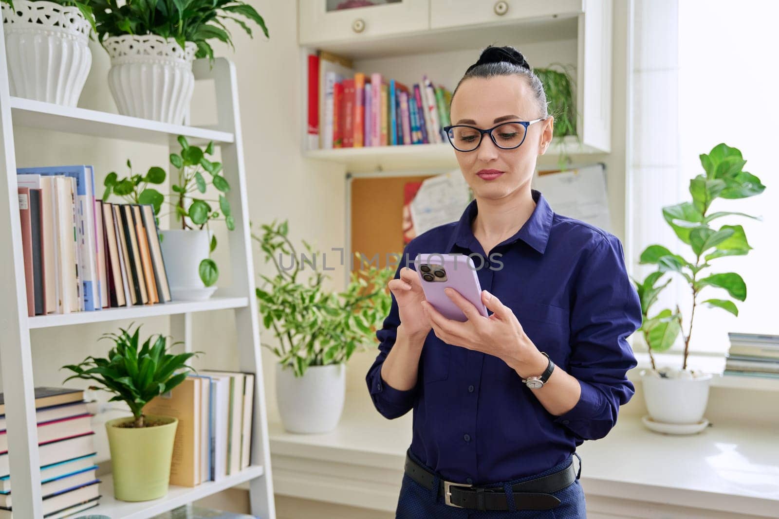 Beautiful serious woman in her 30s using smartphone, standing in her room at home. Communication, work, leisure, lifestyle, people concept