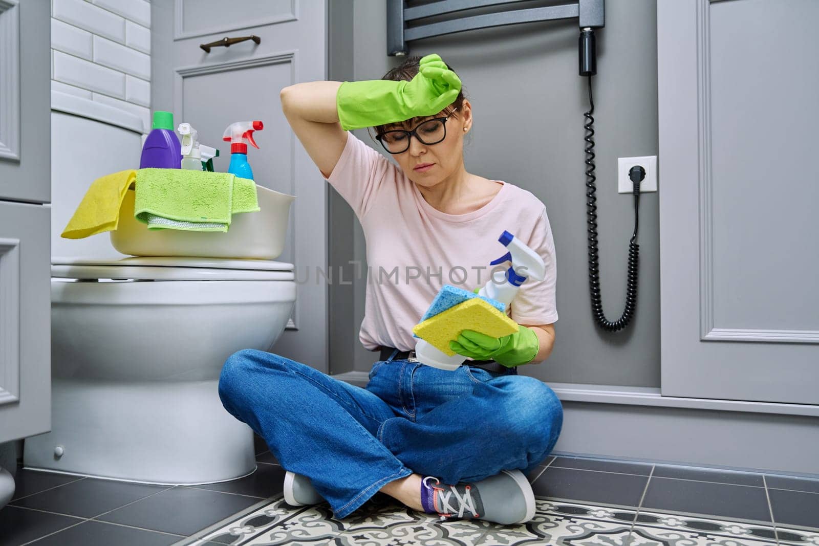 Tired woman sitting on the floor after cleaning bathroom, toilet. Routine house cleaning, home hygiene, housecleaning service, housekeeping, housework