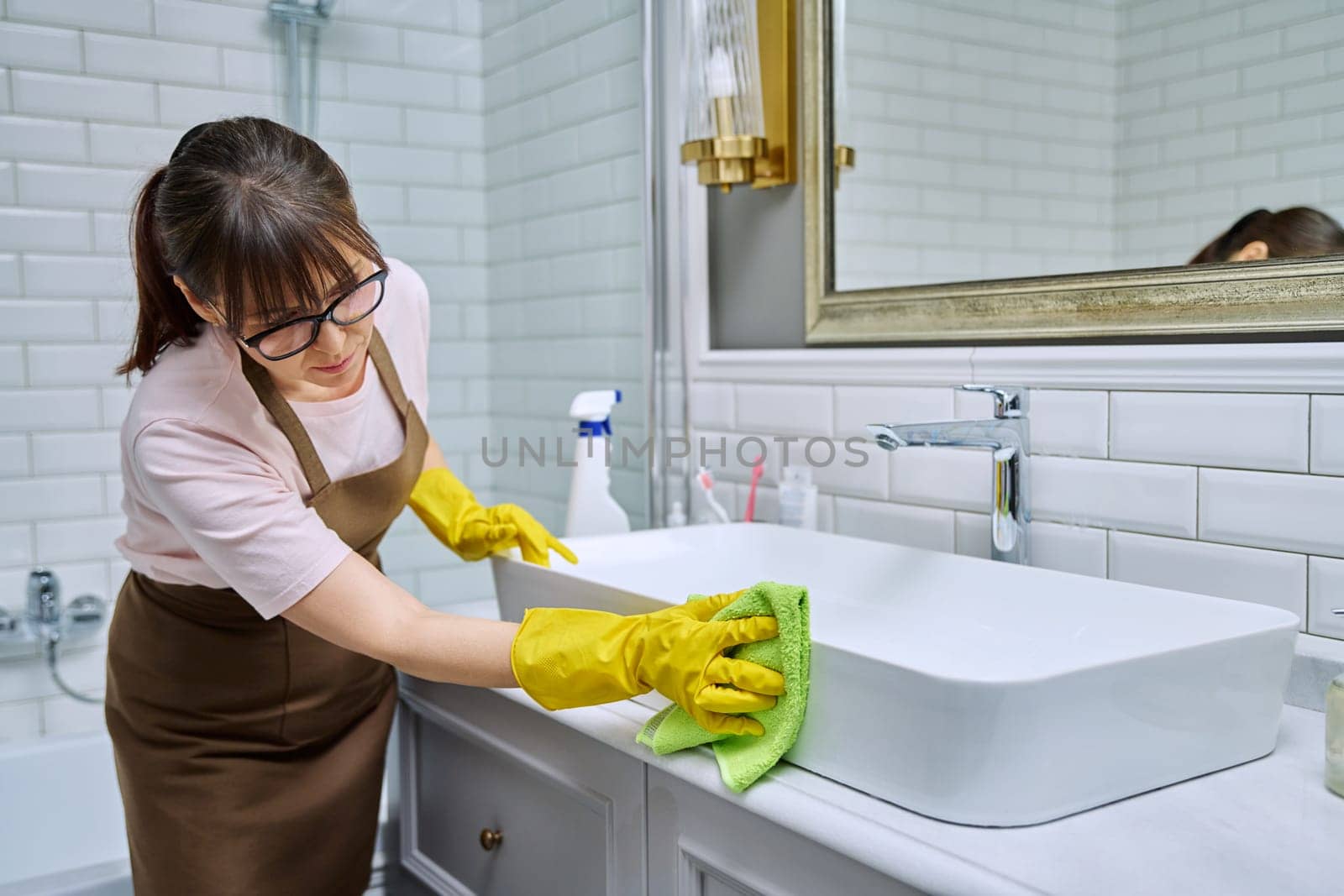 Woman in apron with detergent washcloth cleaning in bathroom, washing sink washbasin. Housewife cleaning house, cleaner service worker at workplace. Home hygiene housecleaning housekeeping housework