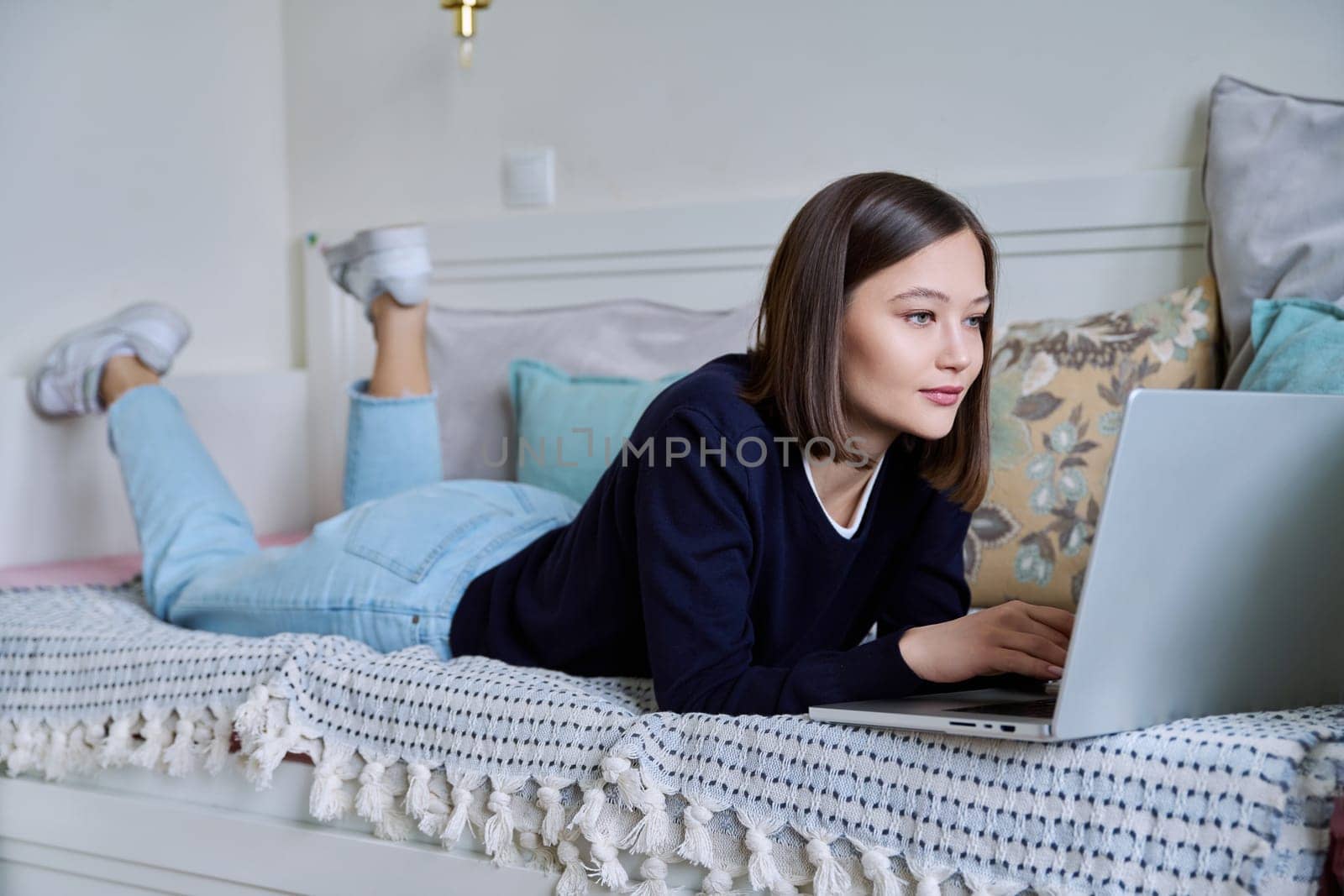 Young woman using laptop computer, typing on keyboard, lying on sofa at home. Internet online technologies for work communication study leisure