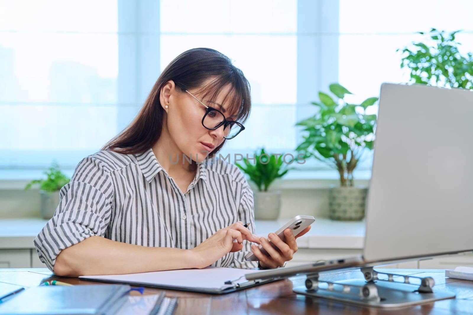 Middle aged woman working with computer papers, using smartphone, remote workplace in home office. Mature people, business, teaching, work, online mental therapy service, freelancing concept