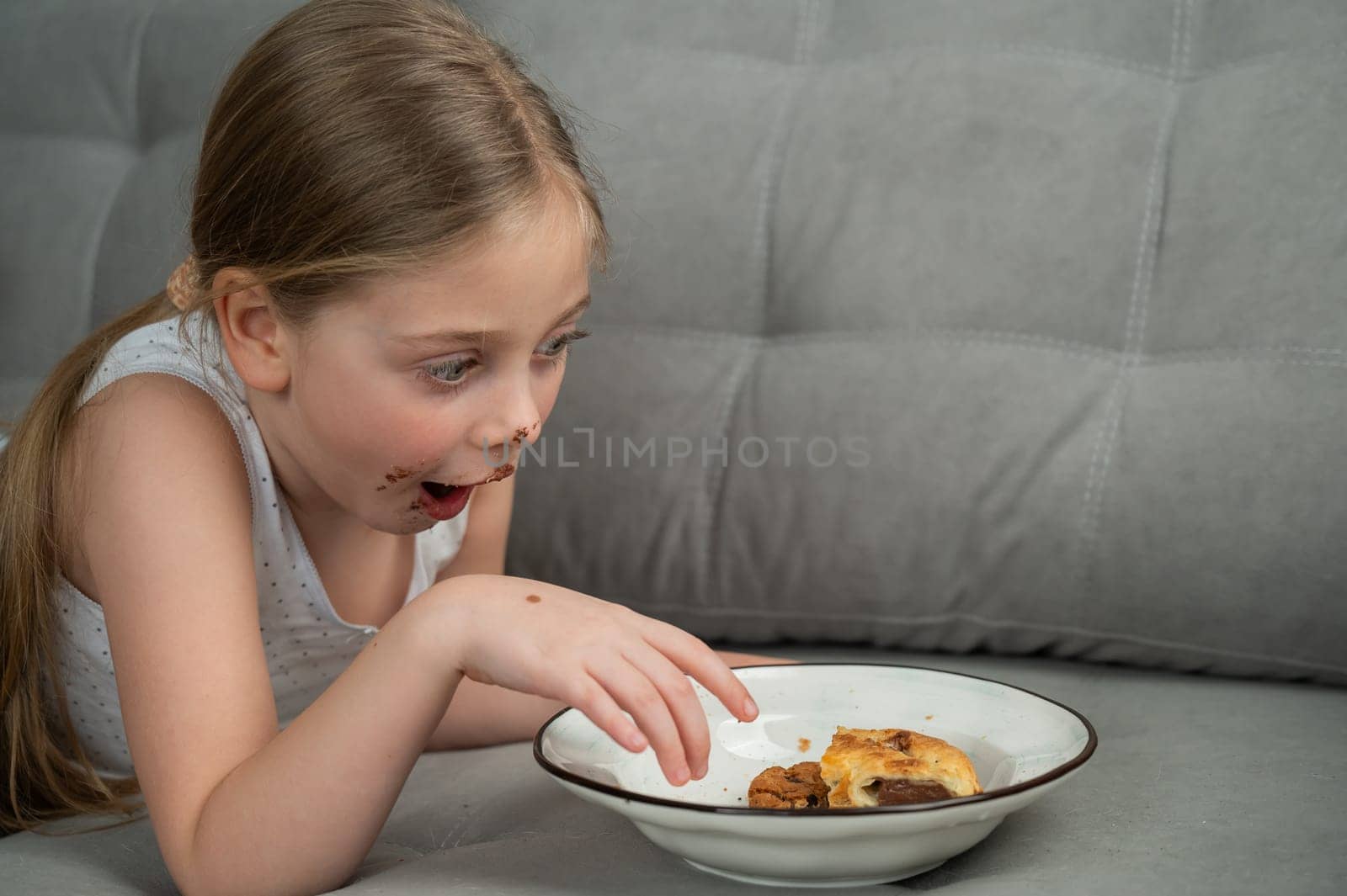 A cute little girl smeared in chocolate eats cookies while lying on the sofa. by mrwed54