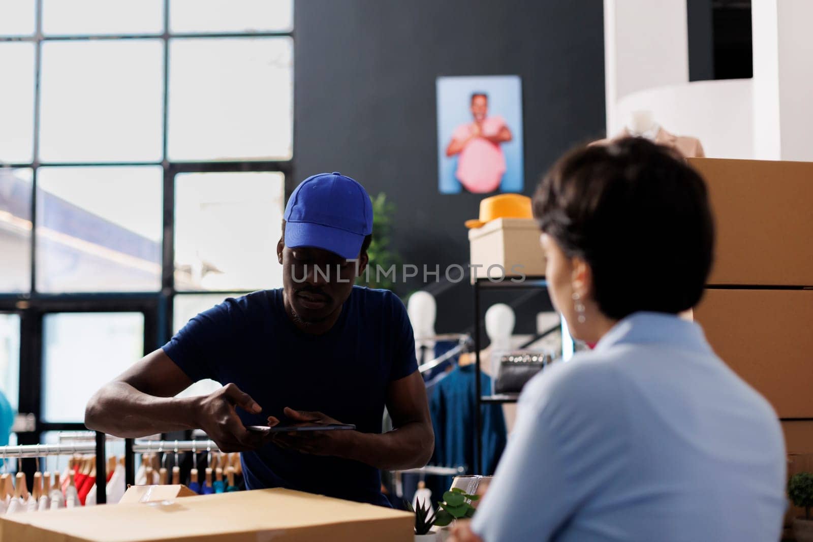 African american courier taking photo of boxes, discussing customer shipping details with employee in clothing store. Worker preparing packages for delivery, working with online orders in boutique