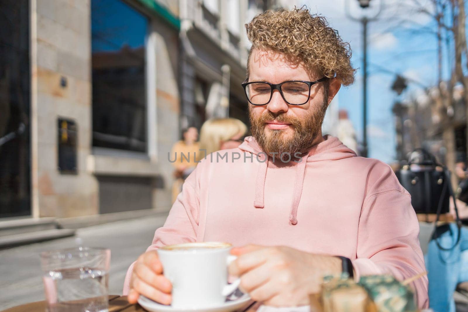 Millennial bearded man having breakfast at table of street cafe on spring day, drinking warm cappuccino. Spring restaurant terrace.