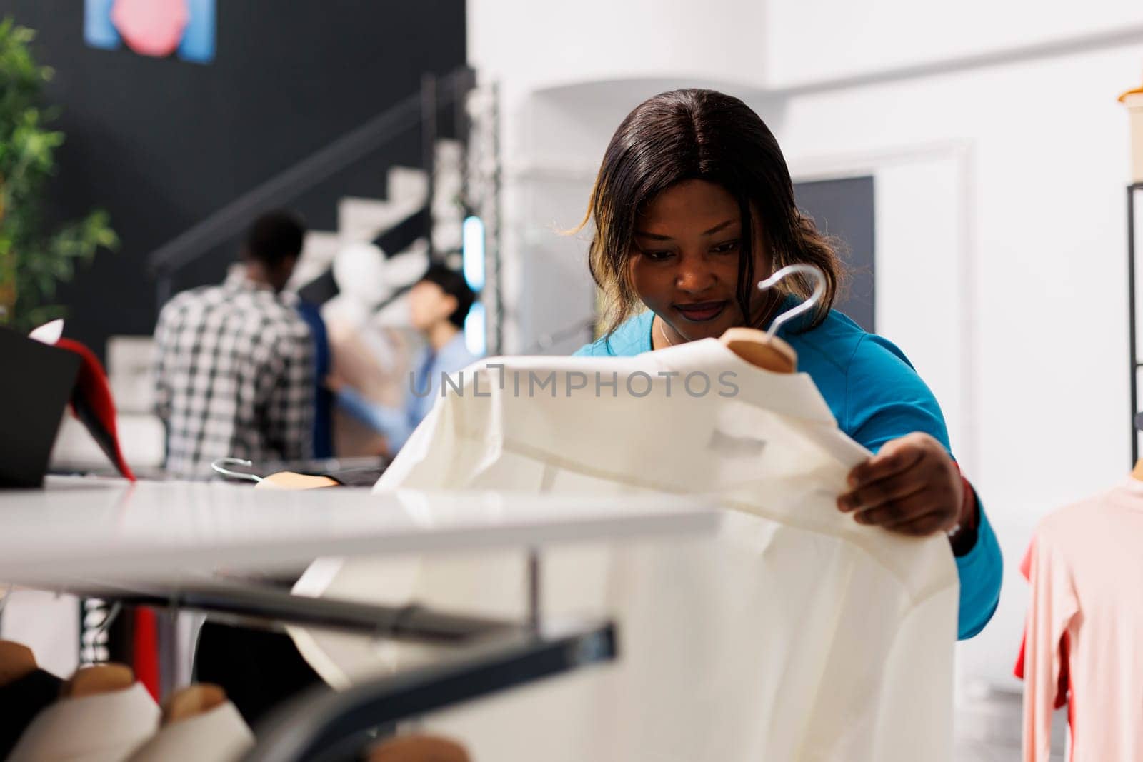 African american customer holding hanger with white shirt, checking items fabric in modern boutique. Shopaholic woman looking at new fashion collection, shopping for casual wear in clothing store