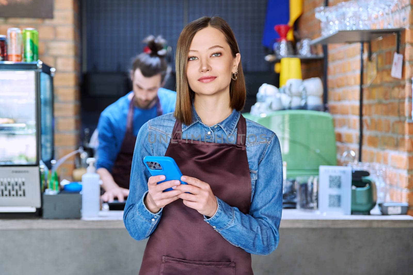 Confident successful young woman service worker in apron holding smartphone in restaurant cafeteria coffee shop pastry shop. Small business, staff, occupation, entrepreneur owner, work concept
