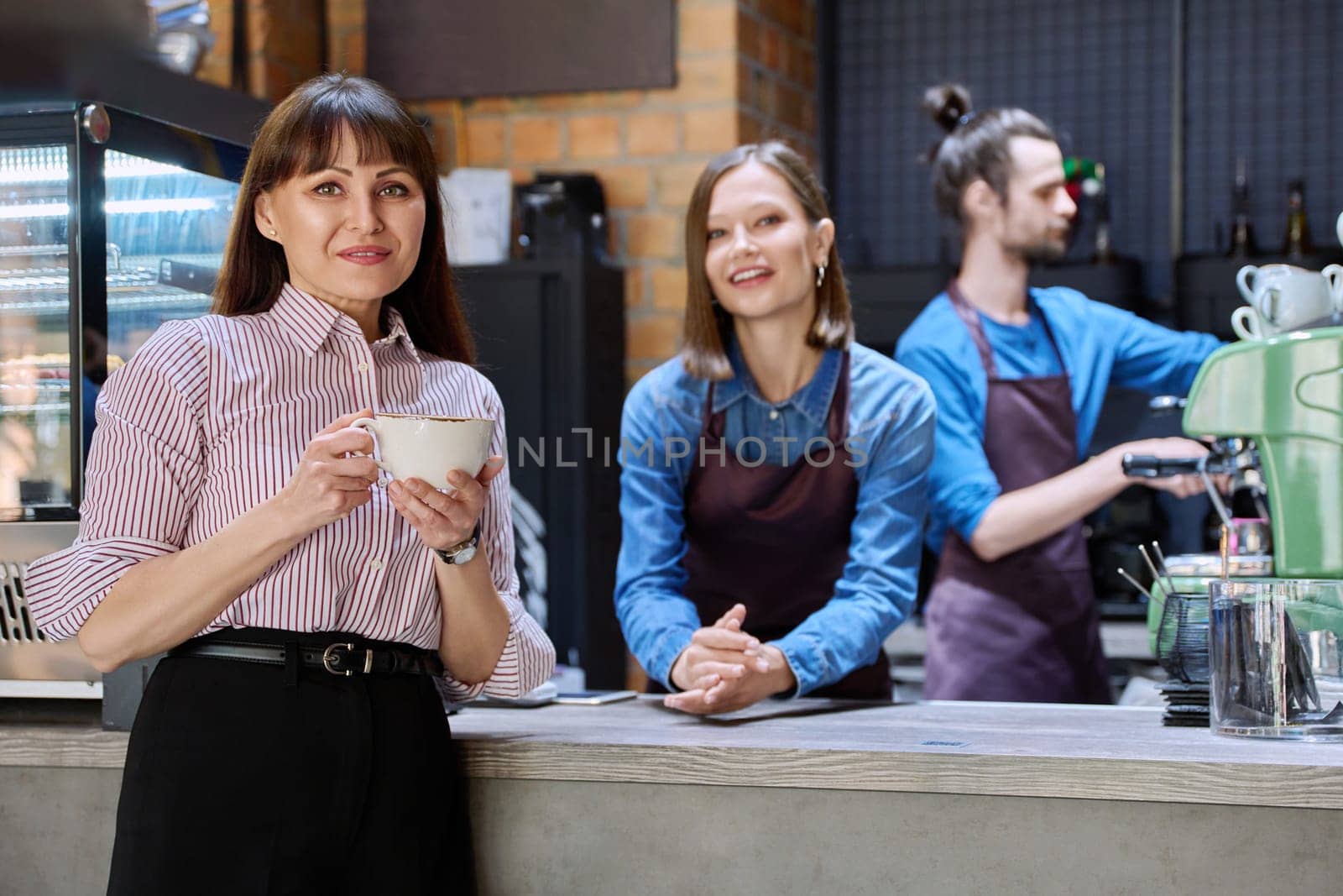 Woman customer of coffee shop near counter with cup of coffee looking at camera, restaurant workers owners at workplace. Colleagues partners in food service work, entrepreneurship, small business