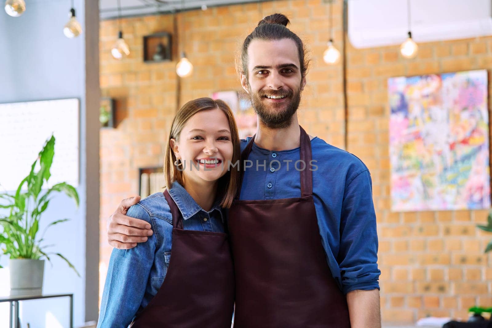 Hugging couple of colleagues partners young man and woman in aprons at workplace by VH-studio